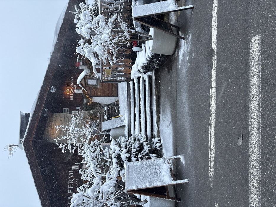 The image depicts the snow-covered exterior of "Hôtel Restaurant Bar La Ferme" in La Clusaz, France, during active snowfall. The building, constructed with dark wood and stone elements, features a steep, dark-toned roof with an antenna. The illuminated sign "HÔTEL RESTAURANT BAR" is visible in orange script on the right side of the facade, while "LA FERME LA CLUSAZ" is visible on the left.
A set of snow-covered steps leads to the main entrance, which is adorned with festive string lights and wall-mounted lanterns. Surrounding the entrance and along the front of the building are numerous evergreen trees and deciduous shrubs, all heavily blanketed with fresh snow. Planters, also covered in snow, contain small trees. A stacked pile of logs, partially obscured by snow, is visible near the steps on the right. Two A-frame chalkboards, completely covered in snow, stand on the pavement in the foreground, one on the left and one on the right.
The asphalt road in the foreground is dusted with light snow, revealing two white painted lines, possibly indicating a pedestrian crossing or parking zone. The ambient light suggests an overcast day. No individuals or active vehicles are present in the scene.