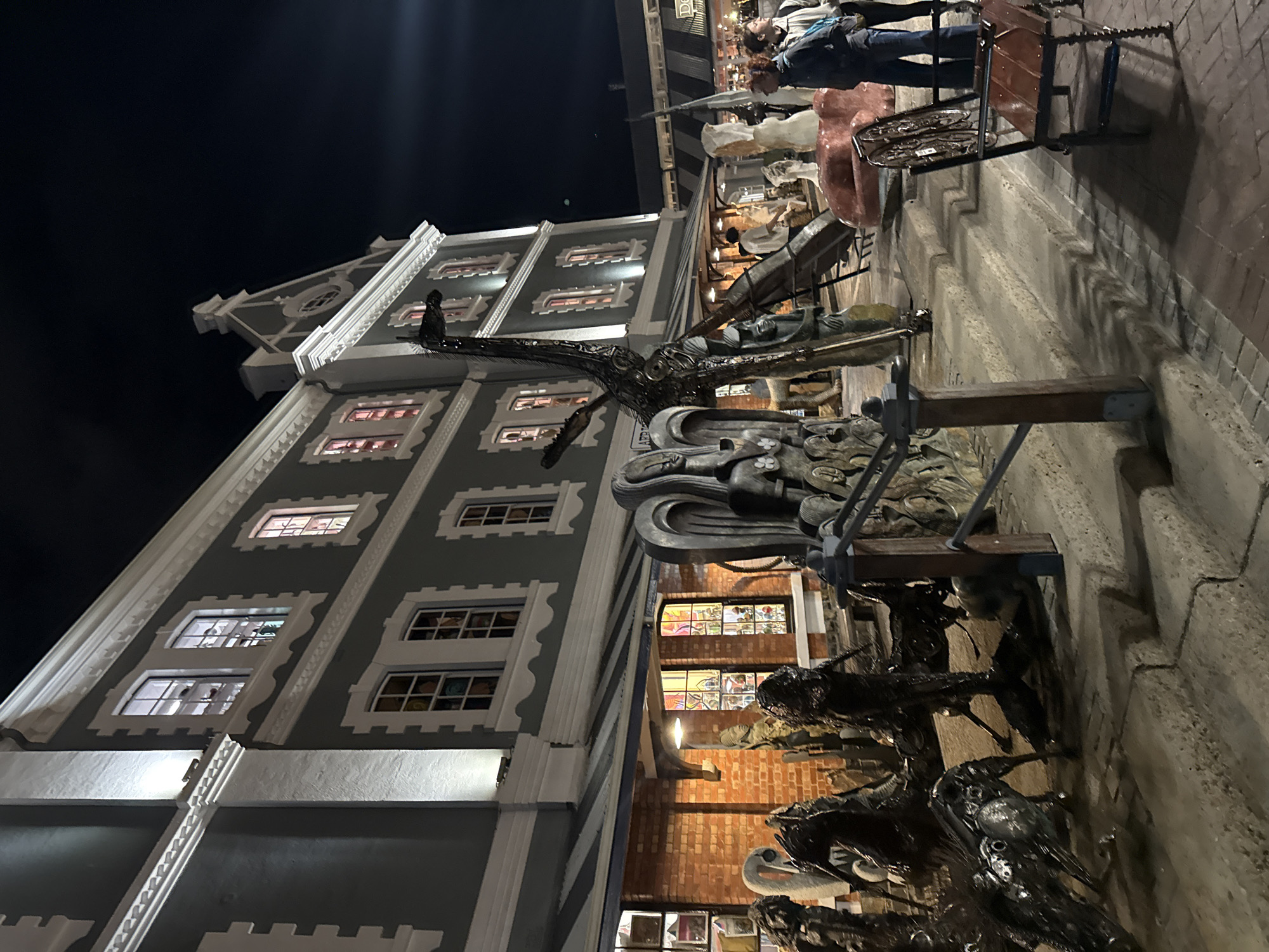 Captured at night in Cape Town, South Africa, the image portrays a blend of architectural and artistic elements. An ornate gray and white building forms the backdrop, its windows lit from within. A large, complex metal sculpture stands in front of it. The sculpture includes depictions of birds. The setting suggests an urban, possibly commercial, area. People are visible near the structure, adding a sense of scale and context to the scene.