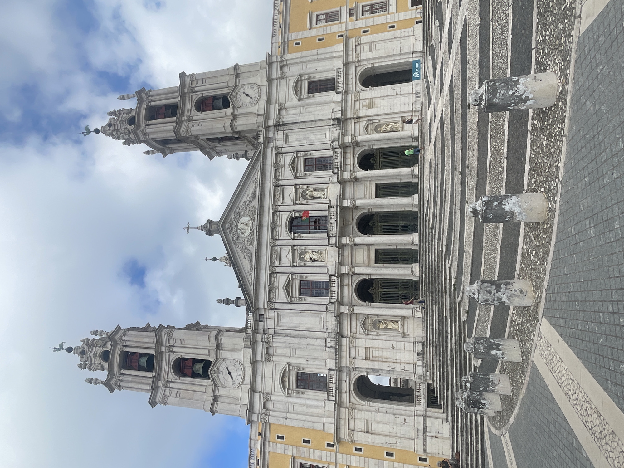 The image depicts the expansive facade of the Mafra National Palace in Mafra, Portugal, under a cloudy sky. The palace features a grand central section flanked by two imposing bell towers. The architecture is characterized by white stone, classical columns, arched doorways, and numerous windows. The foreground of the image shows a large, tiered stone staircase leading up to the palace entrance, with a circular courtyard paved with a mosaic of dark and light stones and cobblestones. Several weathered cylindrical stone bollards are arranged in a curve in the foreground, partially obscuring the lower portion of the steps. The steps themselves are lined with dark bands and feature small decorative stone elements. A few individuals are visible on the steps and in the courtyard, appearing to be tourists. One person is seated on the steps on the left, while others are walking up or standing near the entrance. The overall scene conveys a sense of historical grandeur and scale.