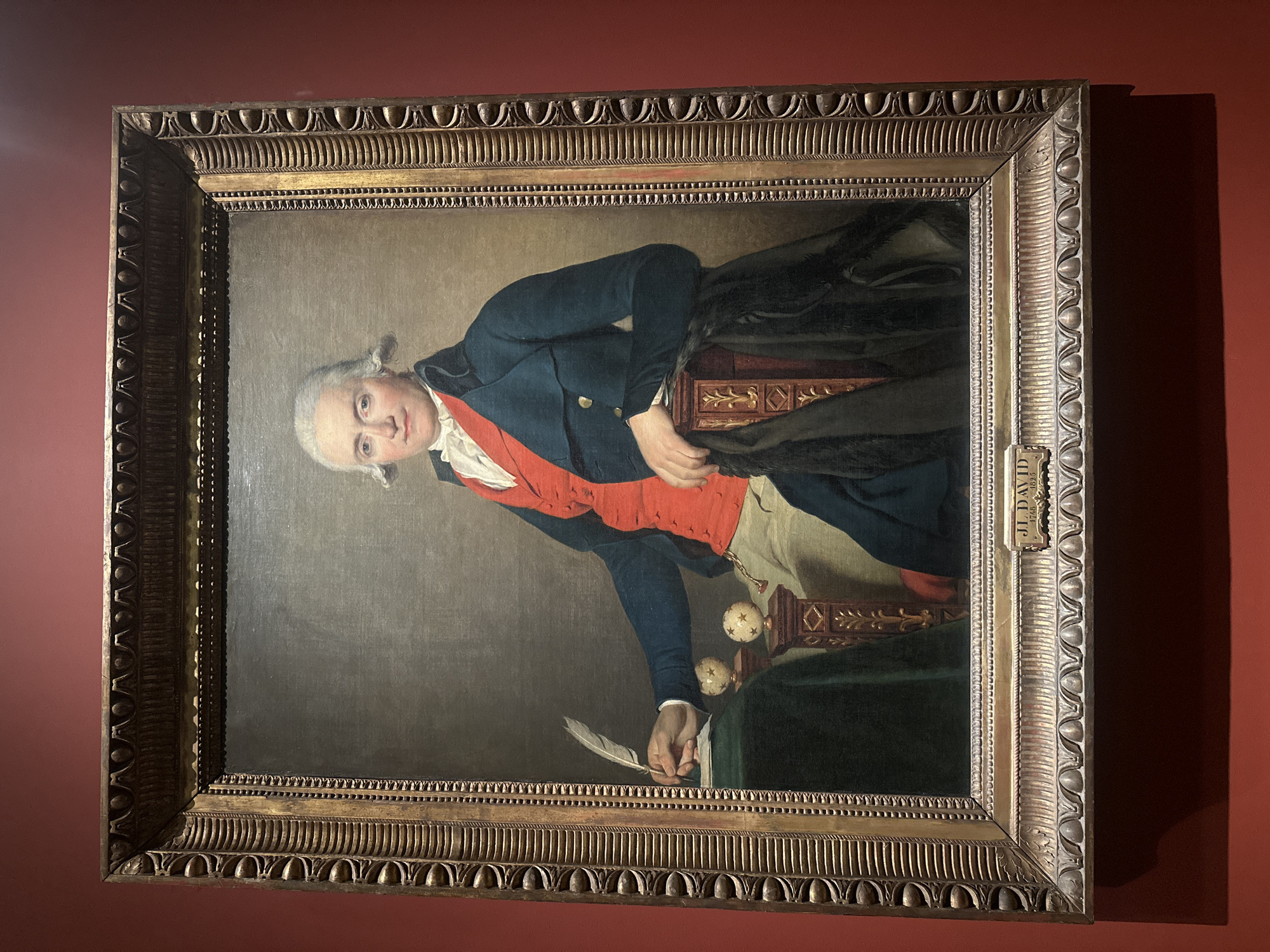 This is a full-length portrait of a man in formal attire, seated and holding a quill. The man has white hair styled in curls and is wearing a white cravat, a red waistcoat, and a dark blue coat. He is seated at a table covered with a dark green cloth, on which rests a piece of paper. Two small, spherical objects adorned with star patterns are placed on pedestals on the table. The man is leaning on an ornate wooden chair back, with a dark cloak draped over it. The painting is housed in a highly decorative gilded frame with intricate carvings. A small plaque below the painting reads "J. L. DAVID 1748 1825".
This artwork is displayed at the Musée du Louvre in Paris, France. The museum is located on the Right Bank of the Seine in the 1st arrondissement of Paris.