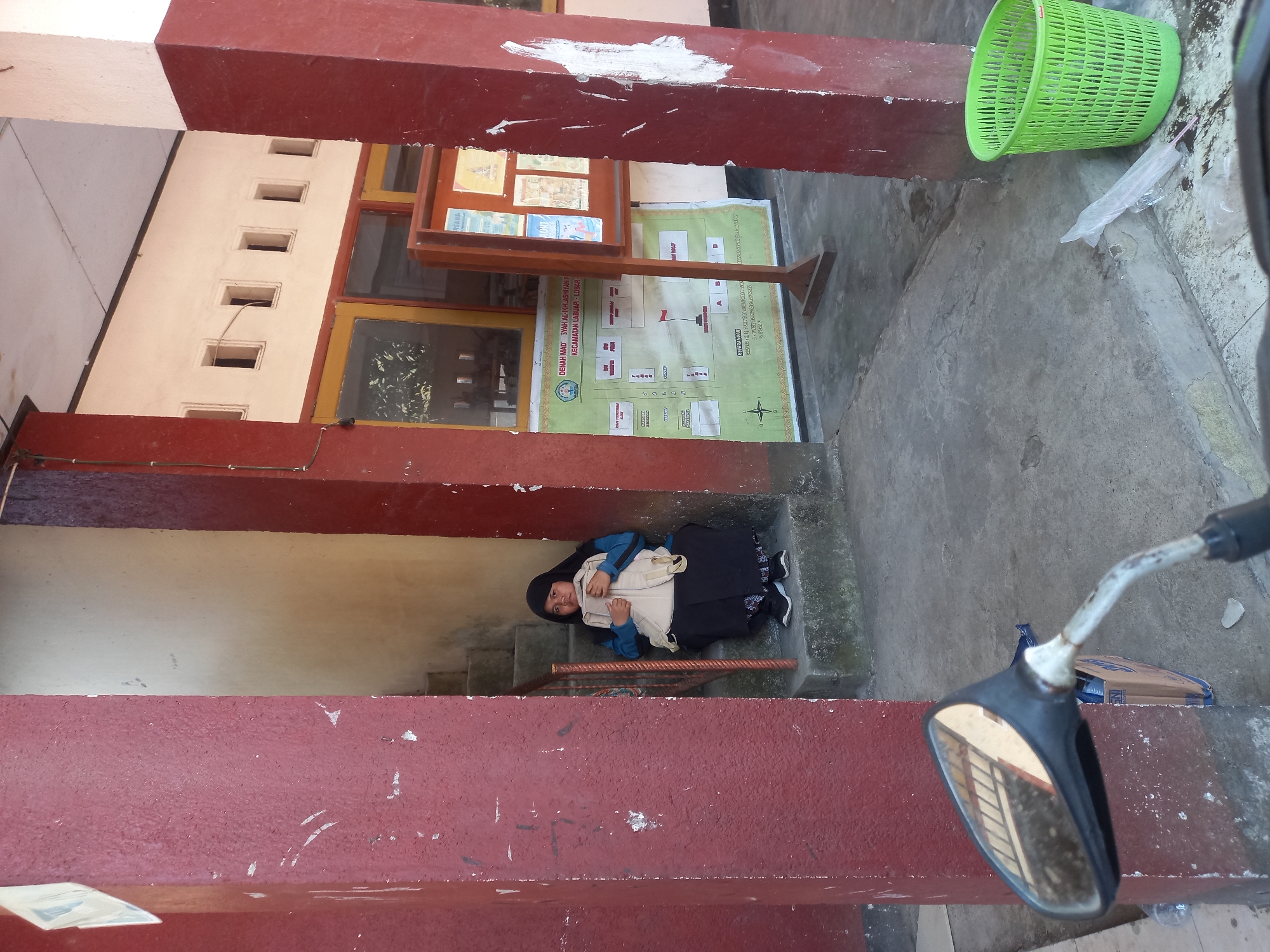 A young girl wearing a black hijab and a blue jacket sits on concrete steps in front of a building. She is holding a beige bag and a light-colored object. The building has a red-painted facade and windows. A green plastic trash can is visible to the right. A motorcycle's rearview mirror is in the foreground, reflecting the stairs. The scene appears to be an outdoor area near a building in Perampuan, Indonesia.