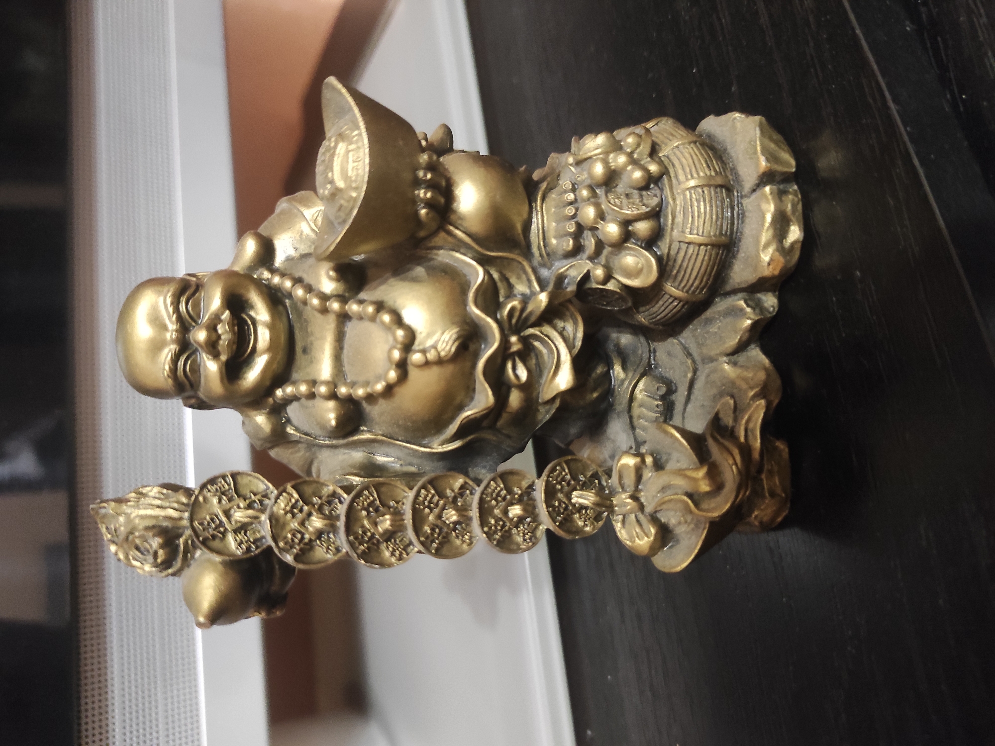 A brass-colored statue of Hotei, or the Laughing Buddha, is the central subject, depicted with a bald head, a portly belly, and a wide, joyful smile. The figure wears a beaded necklace and is adorned with various symbols of wealth and prosperity. These include a large, boat-shaped gold ingot (Yuanbao) near his back, several gourds (Hulu) behind him, and a barrel overflowing with grapes and ancient Chinese coins to his right. A string of interconnected cash coins, each featuring a square hole and unreadable symbols, extends from his left hand. The statue rests on a dark surface, possibly wood, with subtle grain visible. The blurred background reveals an indoor setting, with a light-colored, vertically textured object (perhaps a radiator or blinds) on the left and a warm, muted brown-orange area, likely a wall or furniture, on the right. The lighting suggests an ambient indoor environment. The statue is static, symbolizing abundance and happiness. While intricate designs and symbols are present on the coins and ingot, no legible text is visible.