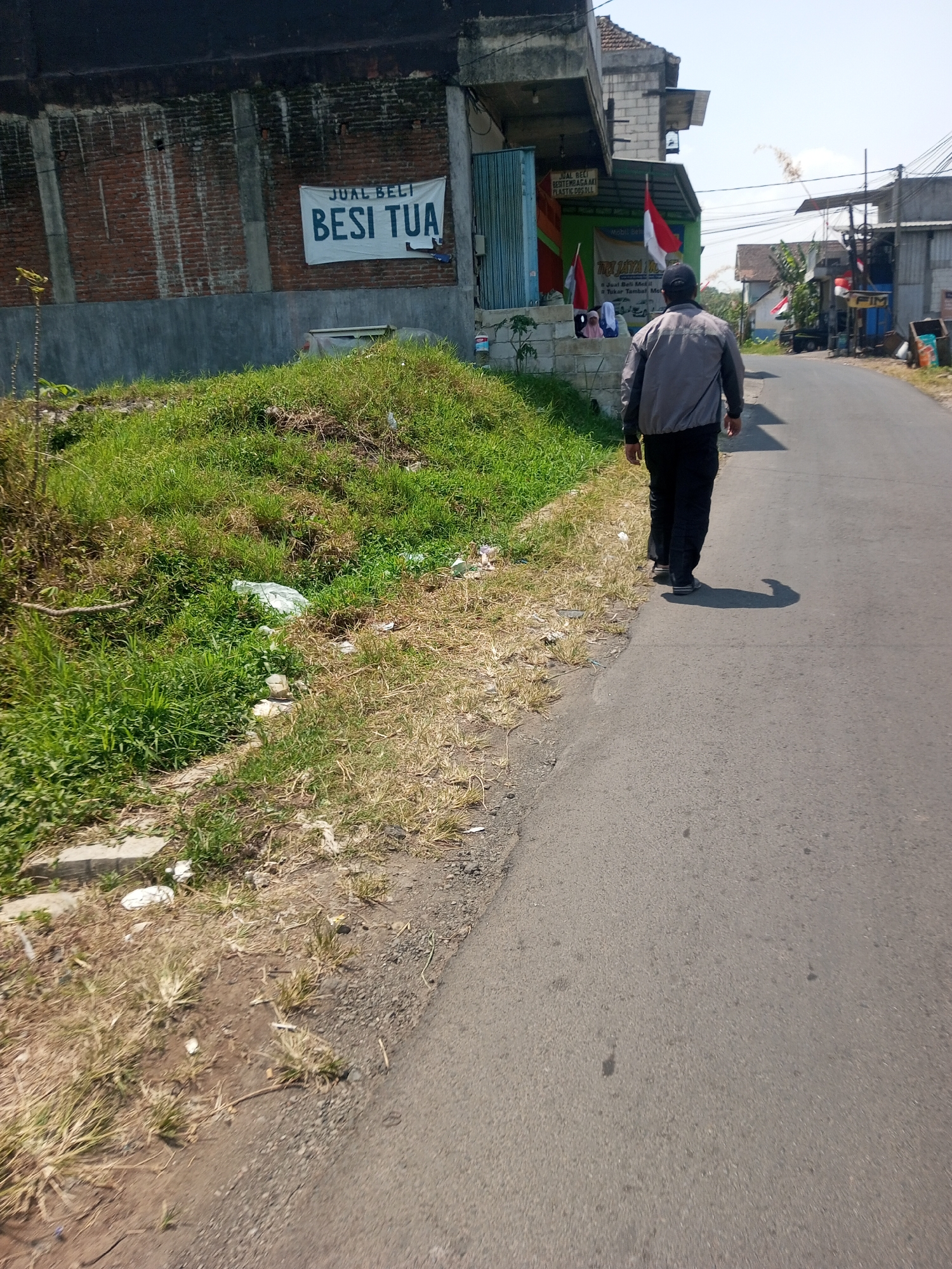 A man walks down a paved road in Singosari, Indonesia. He is wearing a gray jacket and black pants, with a dark baseball cap. The road is lined with vegetation on the left side, with a brick building behind it. A white banner on the building reads "JUAL BELI BESI TUA," which translates to "Buying and Selling Old Iron." The right side of the road features more buildings and houses. The sky is clear and blue, indicating a sunny day. The man's shadow is cast on the road behind him.