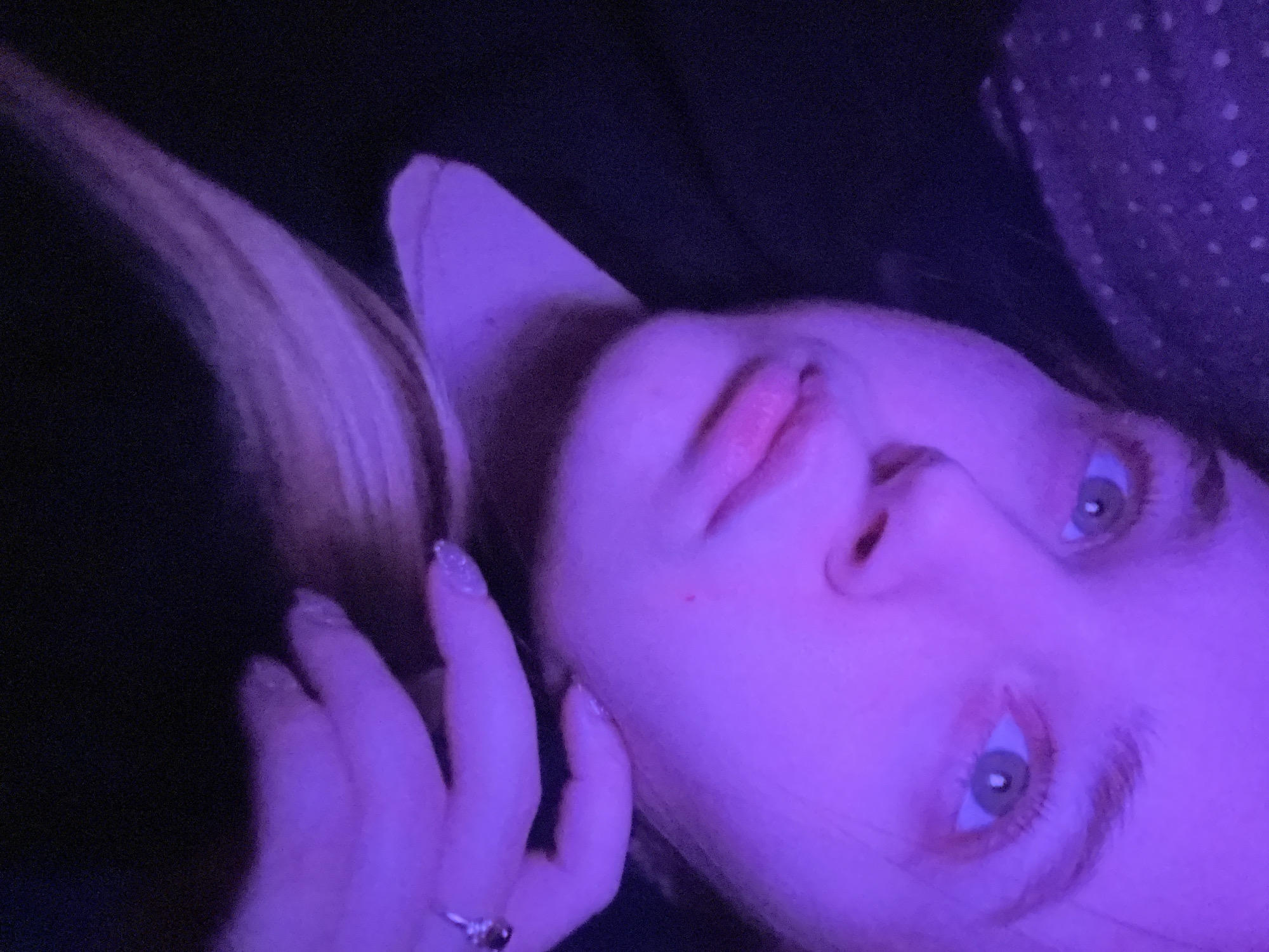 The image displays a close-up, angled view of a young female subject within an indoor setting illuminated by a prominent purple light source. The subject has light-colored eyes, visible pupils, and light eyebrows. Her lips are slightly parted, and a small red mark is visible on her right cheek. Light-colored hair, possibly blonde, extends over her right shoulder. She is wearing a dark garment. Her right hand is raised to the side of her face near her ear, with fingers slightly curled. A silver-toned ring with a dark stone is present on her right ring finger, and her fingernails display a glittery, textured polish. A dark fabric with white polka dots is partially visible on the far left. The background is largely indistinct, appearing dark due to low light. The subject's posture suggests she is reclining or lying down, looking towards the camera. The specific location within Pushchino, Russia, cannot be identified from the visual characteristics present in the image.