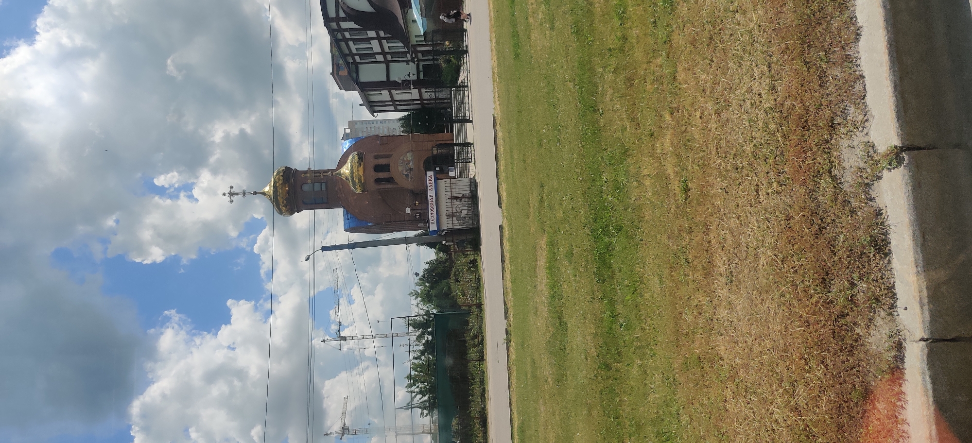 The image captures a view of the Church of the Iberian Icon of the Mother of God in Kharkiv, Ukraine. The church is a red brick structure with golden domes, topped by crosses. A sign in front of the church reads "ЦЕРКОВНА ЛАВКА" (Church Shop). In the background, several construction cranes are visible, indicating ongoing development or repair work in the vicinity. Adjacent to the church, modern buildings with dark roofs and balconies are present. The foreground features a strip of green grass, with a road and sidewalk separating it from the church complex. The sky is partly cloudy, with large white clouds against a blue backdrop.