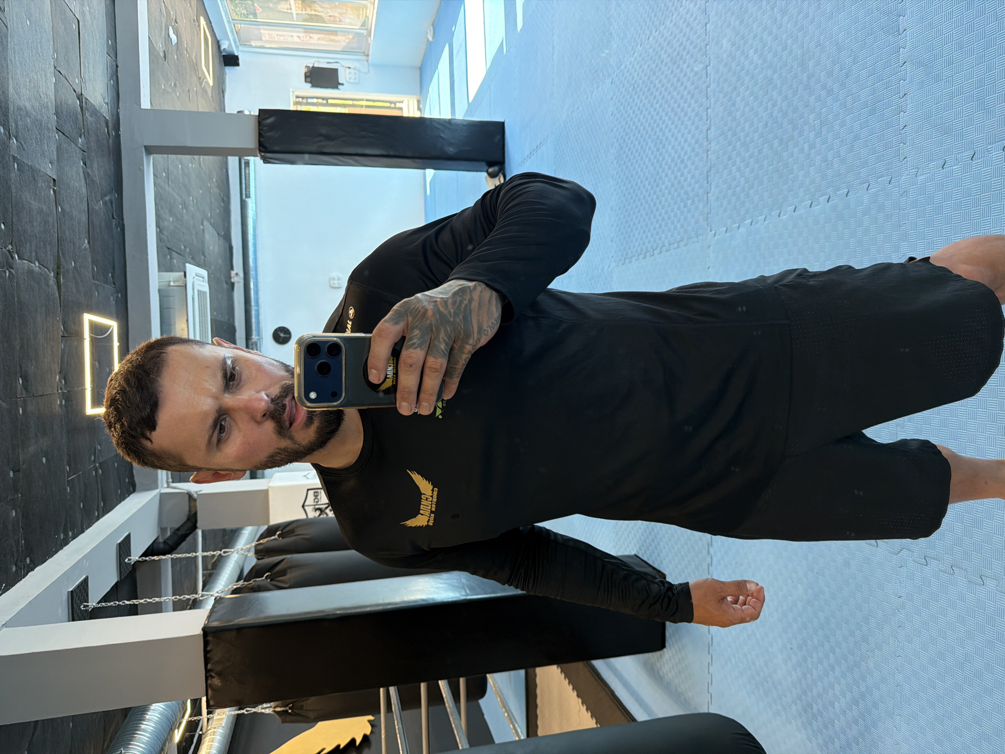 A man with short brown hair and a beard takes a selfie in a gym. He is wearing a black long-sleeved shirt and black shorts. The gym has blue matting on the floor and several black punching bags hanging from the ceiling. In the background, there is a boxing ring and windows that allow natural light to enter the space. The gym appears to be located in Sofia, Bulgaria.