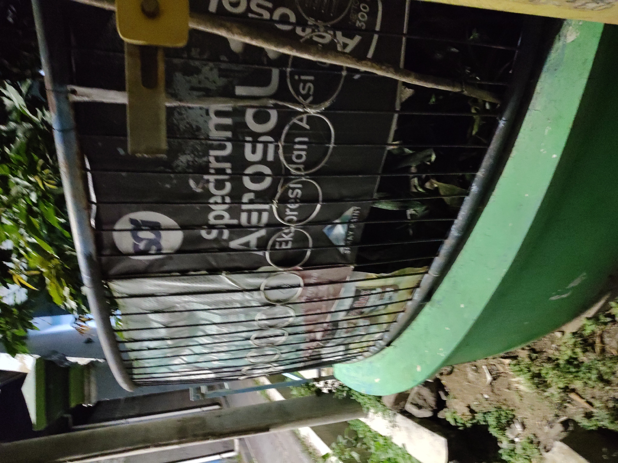 In the city of Unknown, Unknown, a curved green planter box is visible. A black metal fence encloses the planter box, which contains foliage. Behind the fence, on the planter's wall, are several posters. One poster has a white circular logo with a blue "SC" inside. Below this logo, the word "SPECTRUM" is visible in large white letters, followed by "AEROSOL" in even larger white letters. Further down, "Ekspresi dan Aksi" is written in smaller white letters. To the right of these posters, another poster is partially visible with the text "Aerosol" in large white letters and "300" in white below it. A yellow object is attached to the fence, and a wooden pole leans against it. The scene appears to be outdoors at night, with artificial lighting casting shadows.