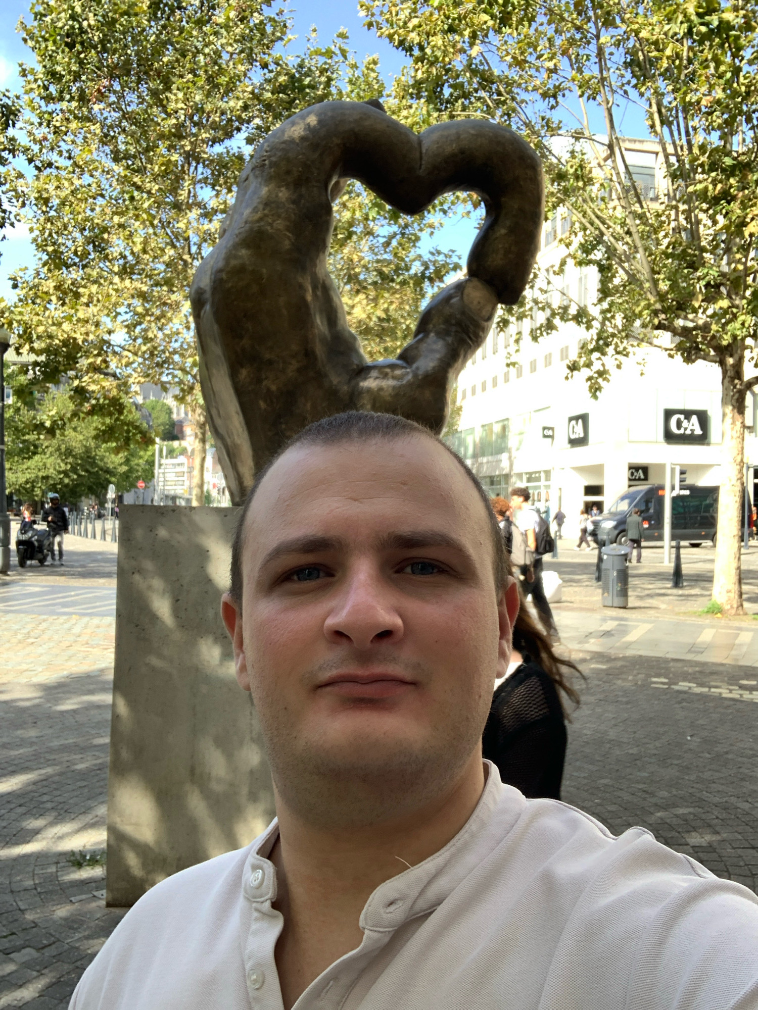 This is a selfie taken outdoors in Liège, Belgium. The subject, a man, is in the foreground, facing the camera. Behind him, a bronze sculpture is visible, which appears to be two hands forming a heart shape, mounted on a concrete base. Trees with green foliage line a paved walkway. In the background, a modern building with the "C&A" logo is visible, along with a dark-colored van and several pedestrians. The street is paved with stone and has streetlights. The sky is blue and clear.