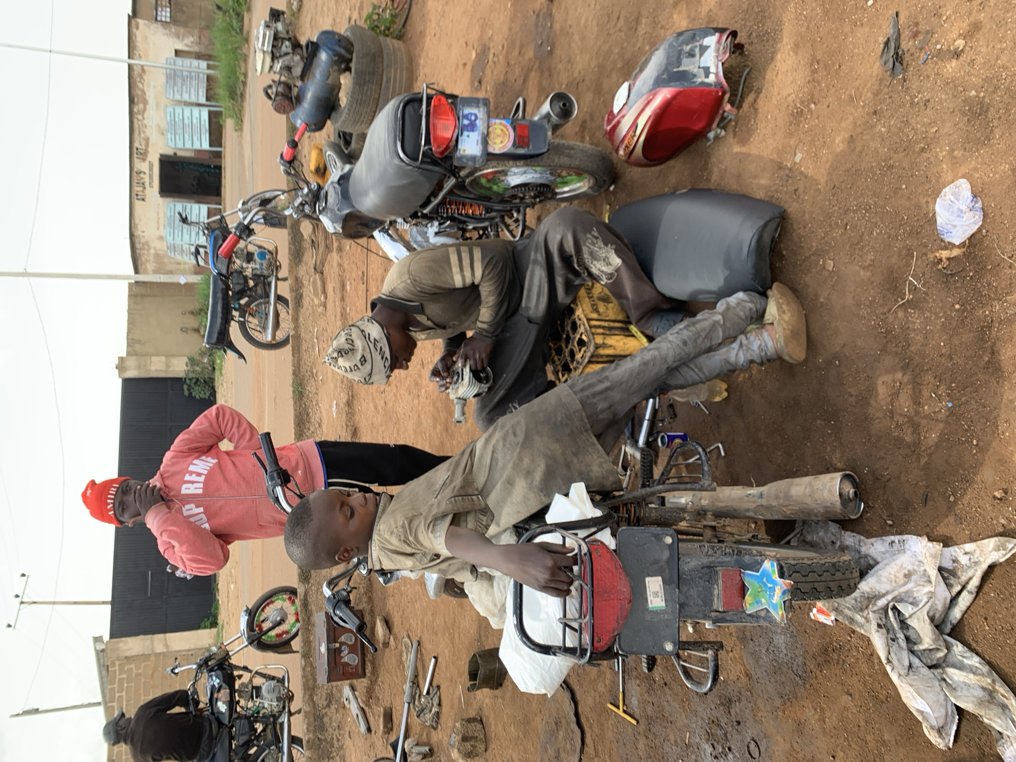 The image depicts an outdoor scene in what appears to be a workshop or roadside repair area in Barakin Lerawi, Nigeria. Two young boys are engaged in repairing motorcycles. The boy in the foreground, wearing a light brown, dusty shirt and pants, is seated on a motorcycle, holding its handlebars. To his right, another boy, wearing a darker shirt and a patterned beanie, is actively working on a motorcycle engine, holding tools in his hands.
Several motorcycles are present in the scene, some partially dismantled. Tools and engine parts are scattered on the dusty ground. An air compressor, a red fuel tank from a motorcycle, and various other mechanical components are visible. In the background, a building with signage that reads "ATAJAY'S ART" is visible. The environment is characterized by reddish-brown earth, suggesting a dry, possibly rural or semi-urban setting. A man in a pink hooded sweatshirt and a red beanie is standing in the mid-ground, observing the activity. Other individuals and motorcycles are also present in the background, indicating a busy workshop environment. The overall atmosphere suggests a functional space for motorcycle repair.