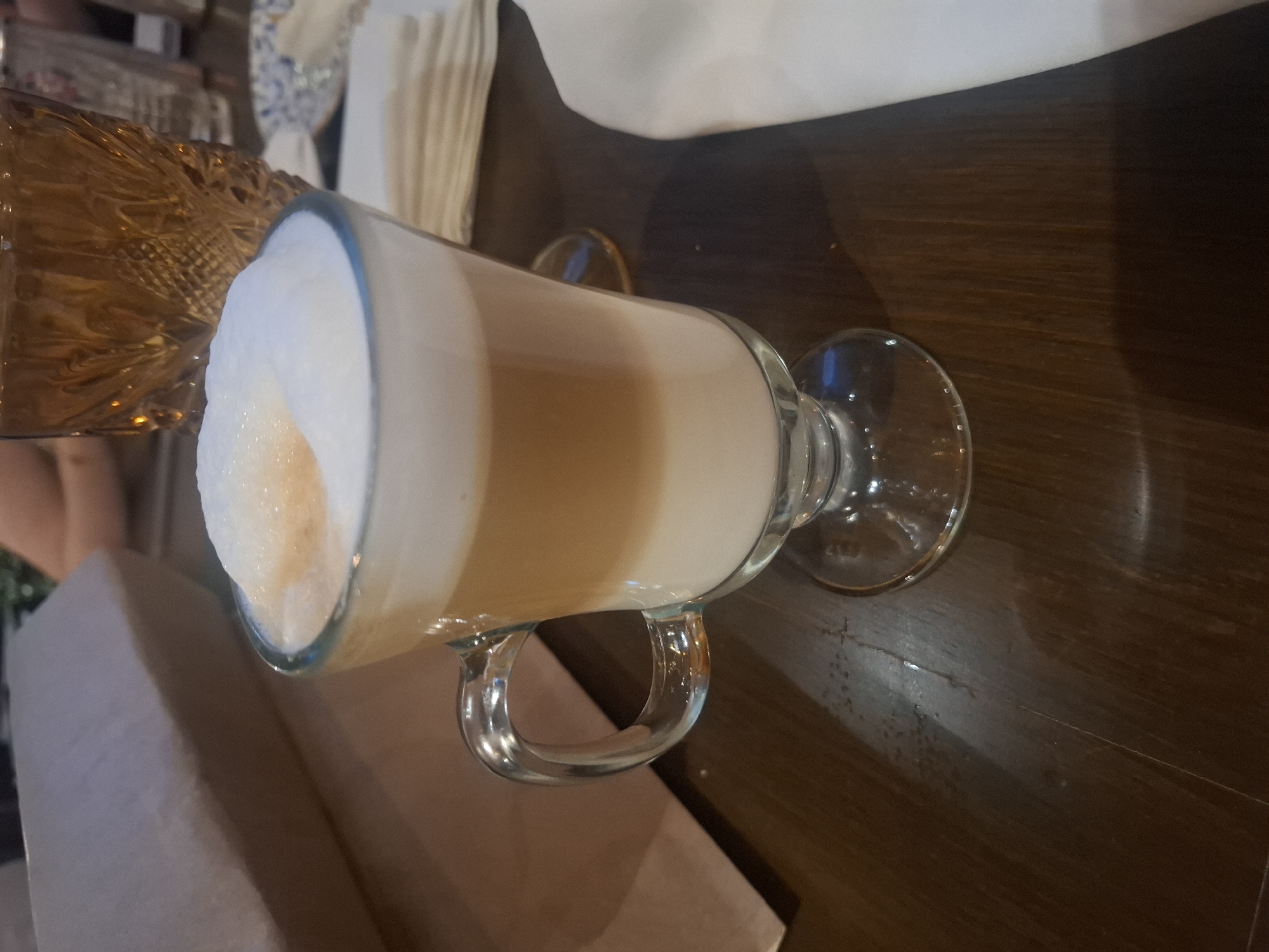 A latte with a thick layer of foam is on a dark wooden table. A decorative glass pitcher sits beside it. Jesús del Monte, Mexico is not discernible in this image.
