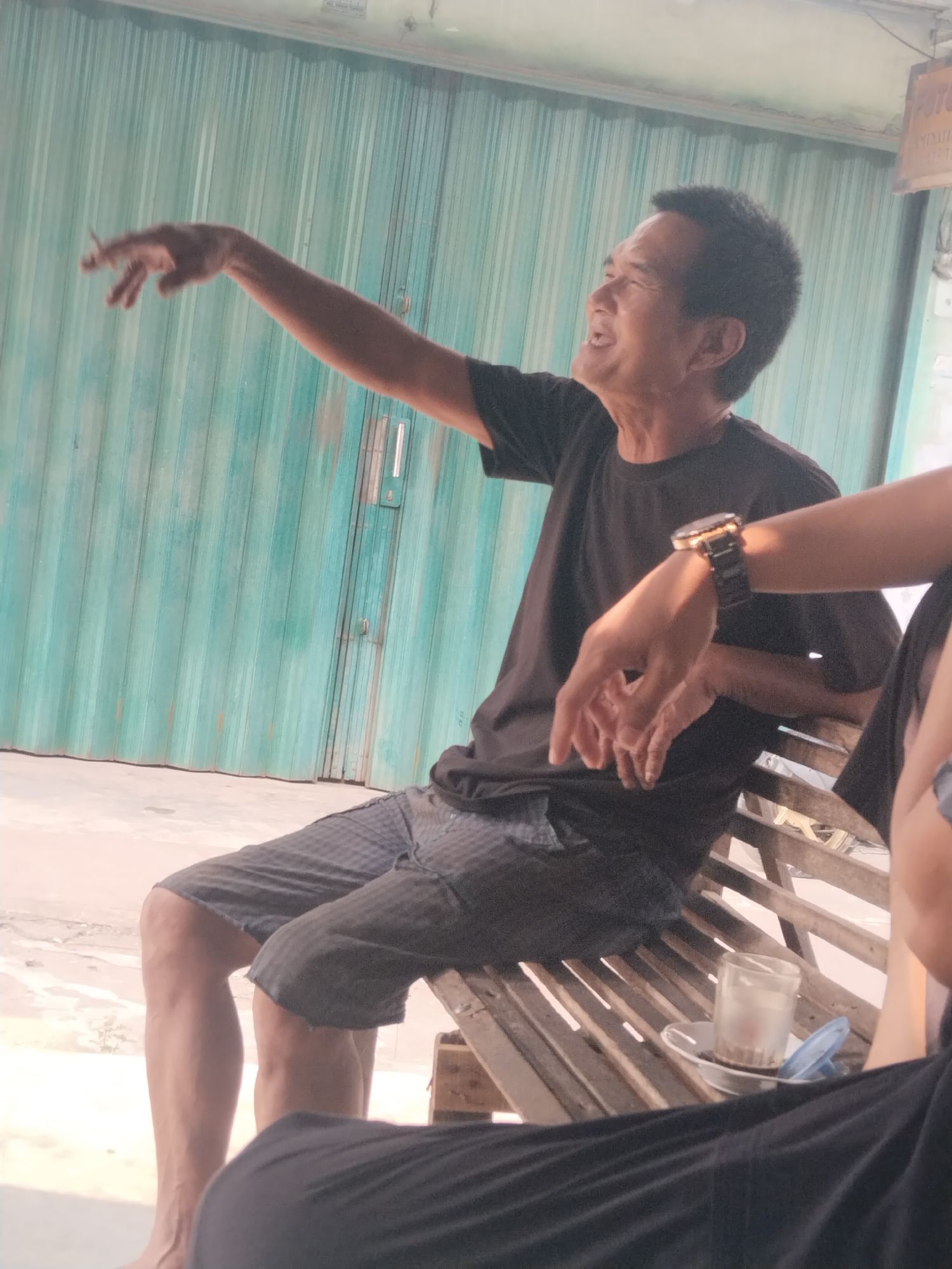 An outdoor scene in Unknown, Indonesia features a man in a black t-shirt and checkered shorts sitting on a wooden slatted bench. He is gesturing with his right hand and appears to be speaking or laughing. A portion of another person's arm, wearing a watch, is visible on the right side of the frame. A small glass with a dark liquid and a saucer are on the bench between the two individuals. The background consists of a light-colored wall and a teal-colored corrugated metal gate or door. The lighting suggests it is daytime.