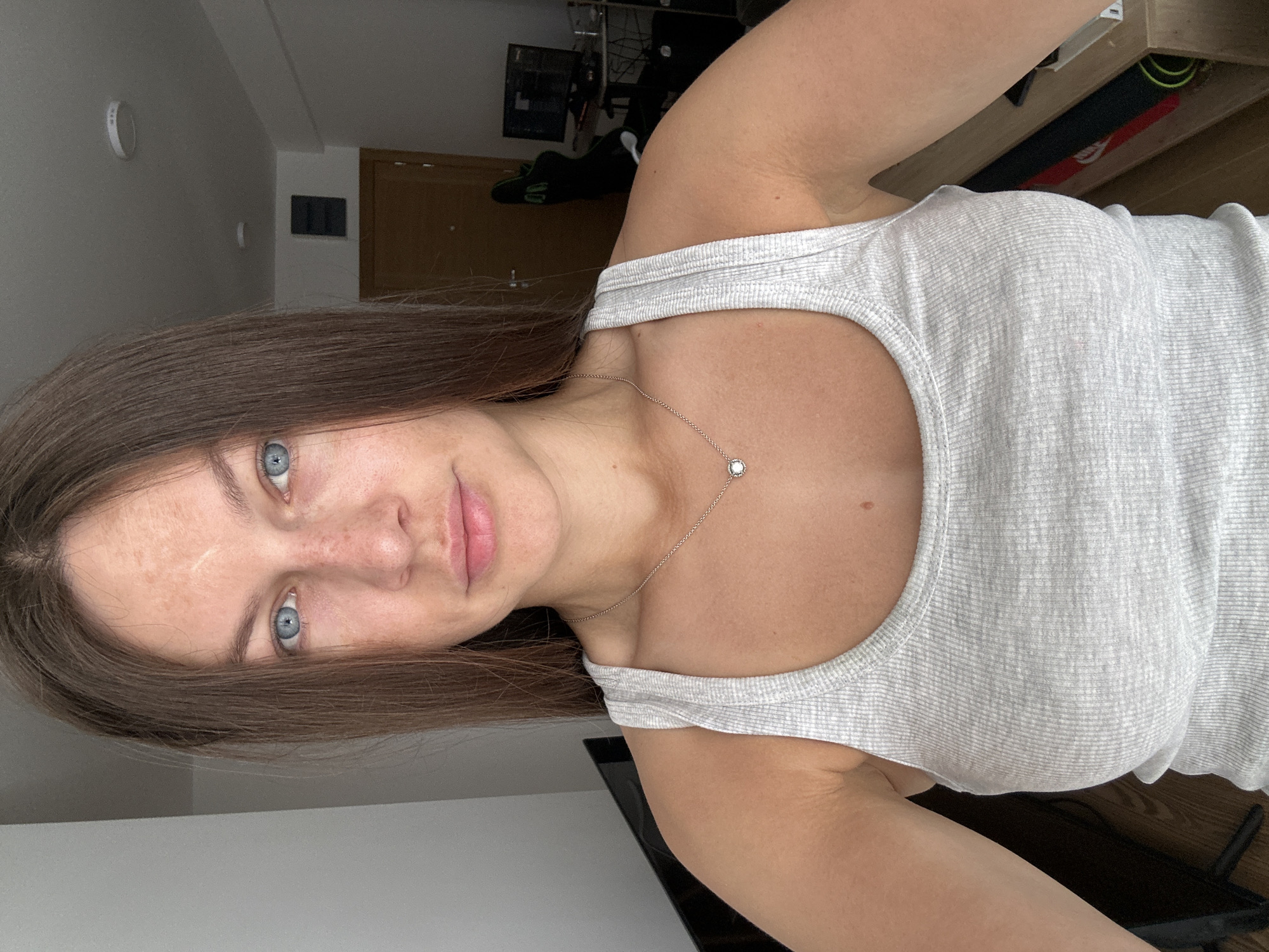 A woman with light skin, light blue eyes, and medium-brown hair is centered in the image. She is wearing a light grey ribbed tank top and a delicate silver necklace with a round pendant. She has a scar on her forehead. The background is a domestic interior, with a door and a wall-mounted control panel visible. A computer setup with a monitor and accessories can be seen in the upper part of the image. The image appears to be taken at a slight angle, giving a portrait-style view. The location is not indicated in the image or implied from any of its elements.