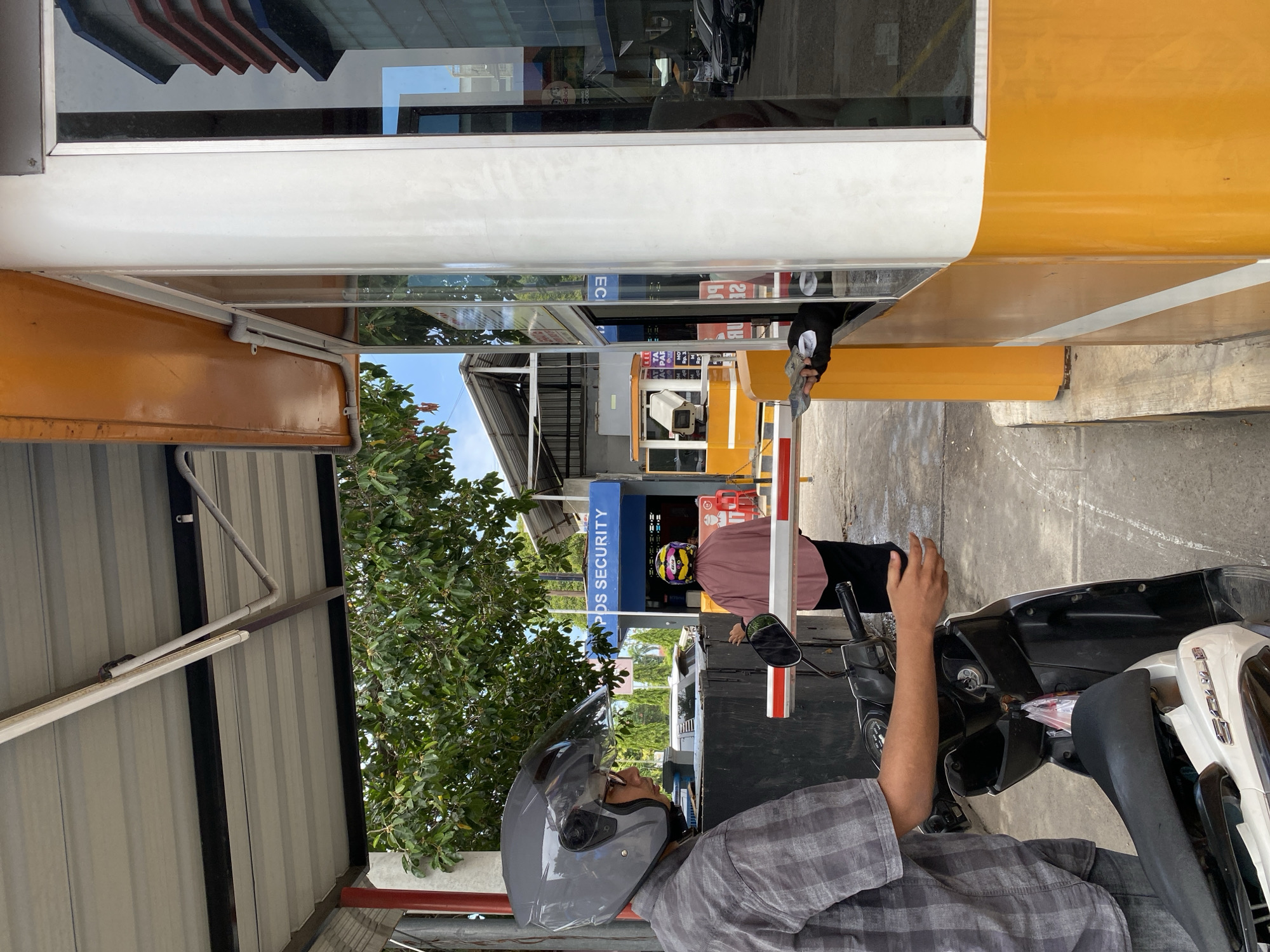 The image depicts an outdoor security checkpoint or entry point in Lhokseumawe, Indonesia.
In the left foreground, a male individual wearing a grey plaid short-sleeved shirt and dark pants is seated on a black and white scooter. He wears a grey full-face helmet with a dark visor partially raised, revealing glasses. His right hand rests on the scooter's handlebar. A red and white striped barrier arm is positioned horizontally in front of the scooter, indicating a controlled access point.
Behind the barrier and slightly to the left, another individual, wearing a pink long-sleeved top and a distinctive yellow helmet with purple and pink markings, is walking towards the background, past the raised barrier. Their right hand is near the barrier mechanism.
To the right, a prominent orange and white security booth features large glass windows that reflect the surroundings, including trees, building structures, and a partial, reversed reflection of the word "SECURITY." A security personnel, dressed in a dark top, extends their hand from an open window of the booth, holding several banknotes and a small white slip of paper, engaging in a transaction. An orange canopy extends from the booth overhead, with white electrical conduit visible underneath.
In the background, a blue building with "PDS SECURITY" prominently displayed in white capital letters identifies the facility. Adjacent to it on the left, a smaller yellow and white booth with a mounted CCTV camera is visible, bearing partially legible signs including "PAR" and "Rp.1," suggesting parking or access fees. Lush green trees with dense foliage fill the mid-ground and background, under a bright sky with scattered white clouds. An overhead corrugated metal roof, supported by a red pillar and black beams, covers the checkpoint area, equipped with fluorescent lighting. The ground is concrete or asphalt.