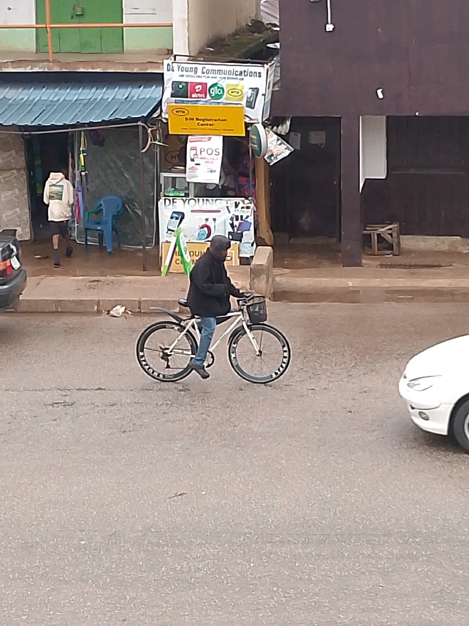 This street-level photograph captures a daytime scene in Bukuru, Nigeria, featuring a man on a bicycle in the foreground. The man is dressed in a dark jacket and light blue jeans, with his feet near the ground as he either rides slowly or is momentarily stopped on a paved road. His bicycle has a white frame, dark wheels with some white text, and a small black basket attached to the handlebars.
In the background, a row of commercial establishments lines the street. On the left, a shop with a blue corrugated metal awning shows a person in a white hooded sweatshirt and dark shorts walking towards its entrance, with a blue plastic chair outside. Adjacent to this, a prominent banner for "Da Young Communications" displays the logos for mobile network providers "airtel," "glo," and "MTN." Below it, a yellow sign advertises "SIM Registyrian Cantor" (likely "SIM Registration Center"), and a "POS" (Point of Sale) sign is also visible. The building to the right features a dark, unadorned facade with a wooden crate or stool placed near its entrance.
On the road, the front section of a white car is visible on the right, and part of a dark-colored car is on the far left, suggesting the presence of vehicles and traffic. The lighting is soft and diffused, indicating an overcast day. The scene depicts a typical busy local street in an urban or semi-urban setting.