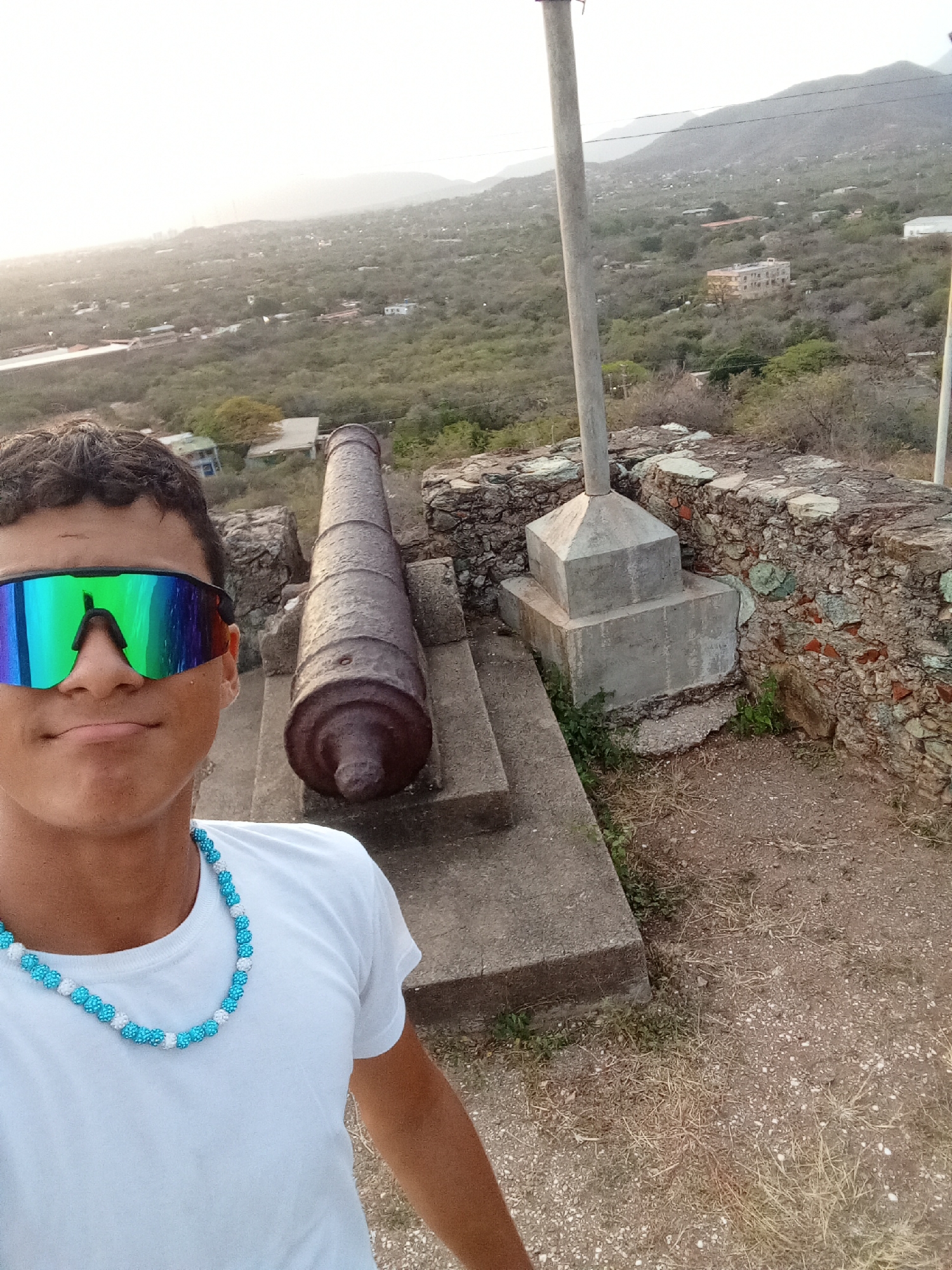 A selfie photograph captures a male subject in the foreground, wearing a white t-shirt, a blue and white beaded necklace, and iridescent sunglasses. Behind him, a weathered, brown cannon rests on a concrete base. A tall, gray pole stands vertically, rooted in a concrete plinth. To the right of the cannon, a rough-hewn stone wall is visible. The background features a densely vegetated landscape with scattered buildings and rolling hills, typical of the topography surrounding the city of Juan Griego, Venezuela. The lighting suggests it is late afternoon or early morning.