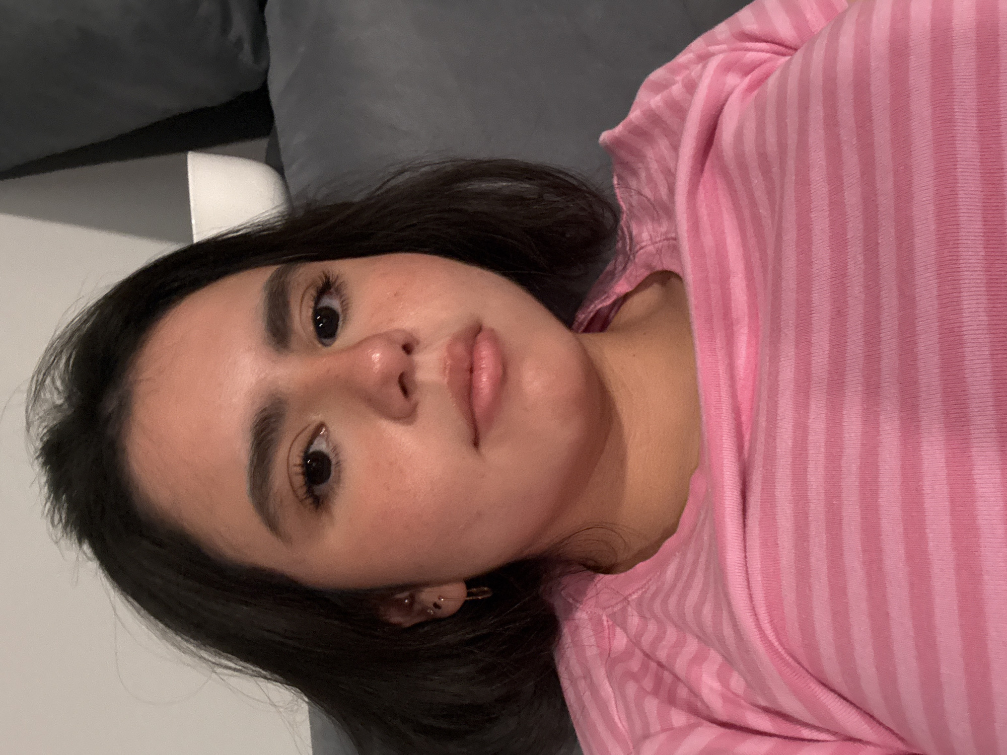 A young woman is shown from the chest up, with her face turned slightly towards the camera. She has dark hair, dark eyes, and appears to be wearing light makeup. She is wearing a pink striped shirt. The background is blurry and appears to be an indoor setting, possibly a couch or sofa. No specific Austin landmarks or context are visible.