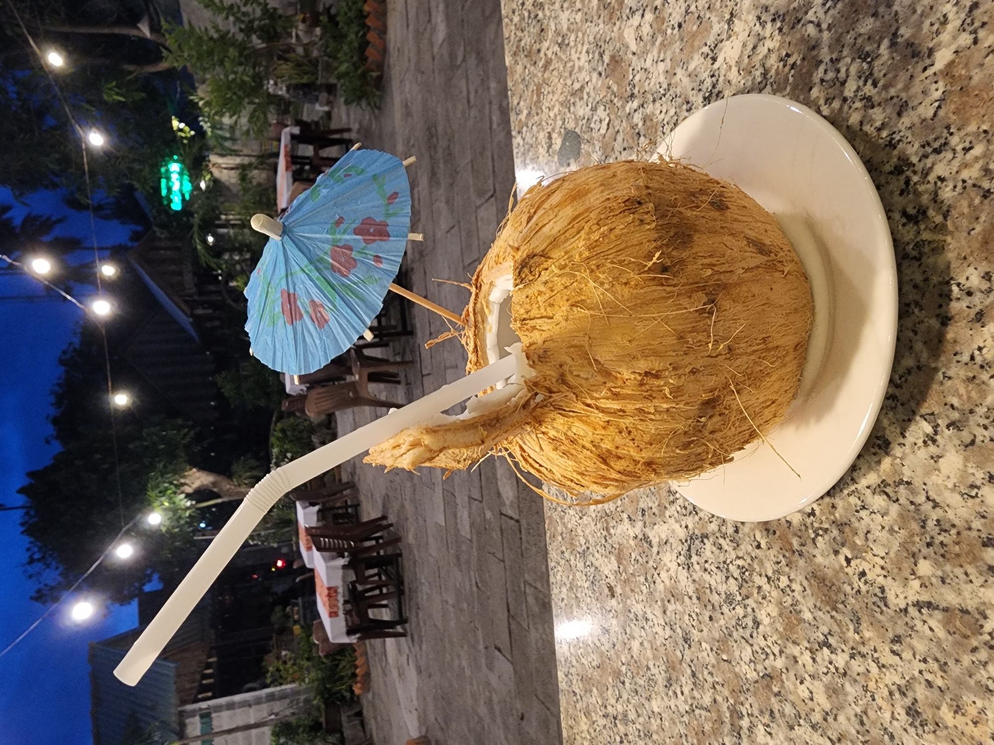 In the foreground, a whole coconut with a straw and a small, blue, floral-patterned umbrella rests on a white plate on a speckled granite countertop. The coconut is hairy and fibrous. In the background, an outdoor seating area with tables and chairs is visible under a twilight sky illuminated by string lights. The setting appears to be a restaurant or resort in Phan Thiet Ward, Vietnam.