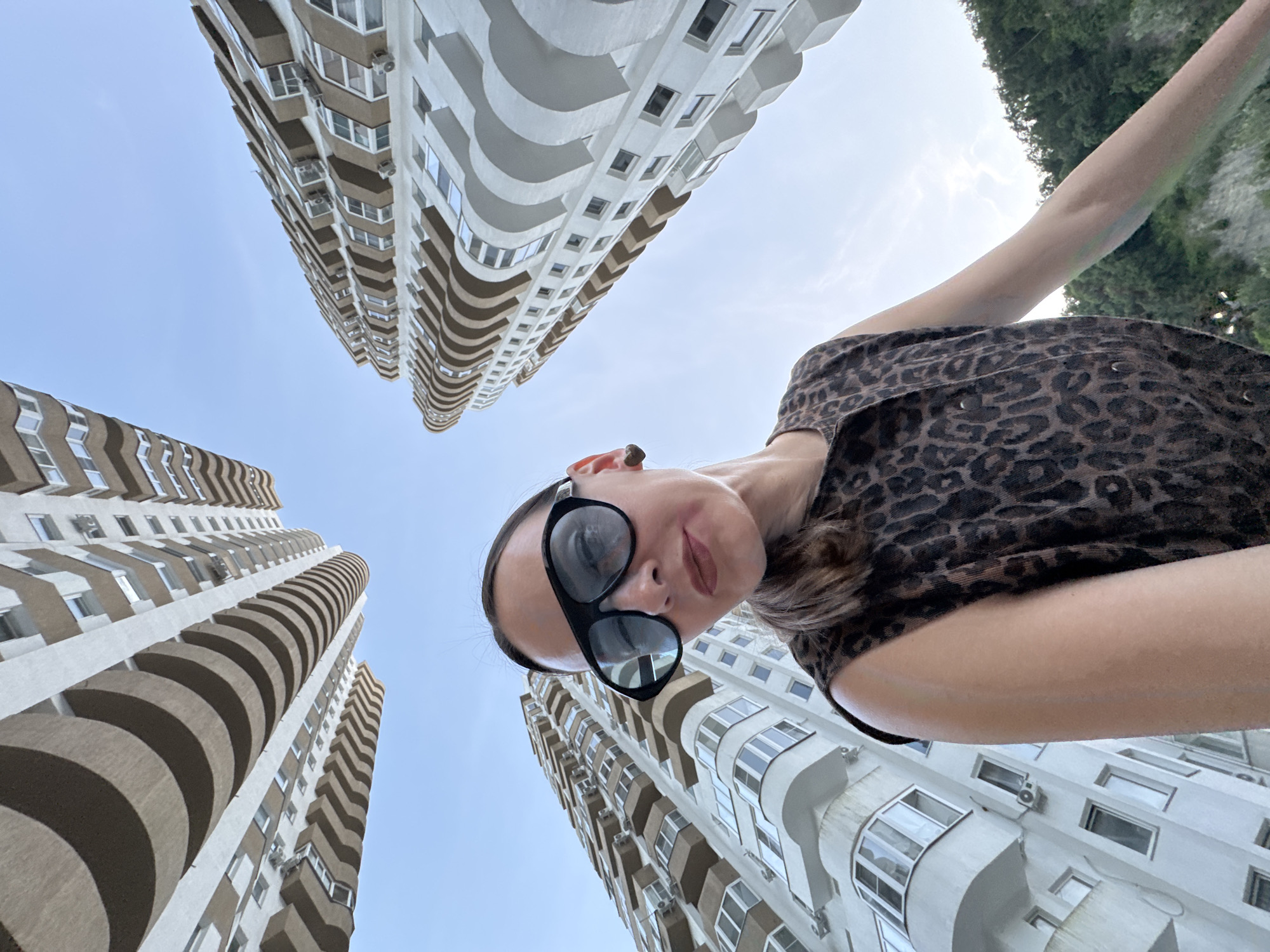 This media file features a low-angle, wide shot looking upwards at a woman taking a selfie. The woman is wearing a leopard-print top and large, black sunglasses. She has her head tilted back, and a slight smile on her face, suggesting a relaxed and possibly playful mood. Her arm is extended upwards, indicating she is holding the camera or phone.
The background is dominated by the architecture of several tall, modern apartment buildings. These buildings are characterized by their beige and white exteriors, featuring curved balconies that create a distinctive, repetitive pattern along their facades. The perspective emphasizes the height and scale of the buildings against a bright, clear sky.
The scene appears to be set outdoors, likely in an urban or suburban environment with a focus on residential architecture. The time of day is likely daytime, given the bright, even lighting and the clear sky. The weather seems fair and pleasant. The context suggests a location in Sochi, Russia, which aligns with the style of modern apartment complexes often found in such areas. No visible text is present in the image.
