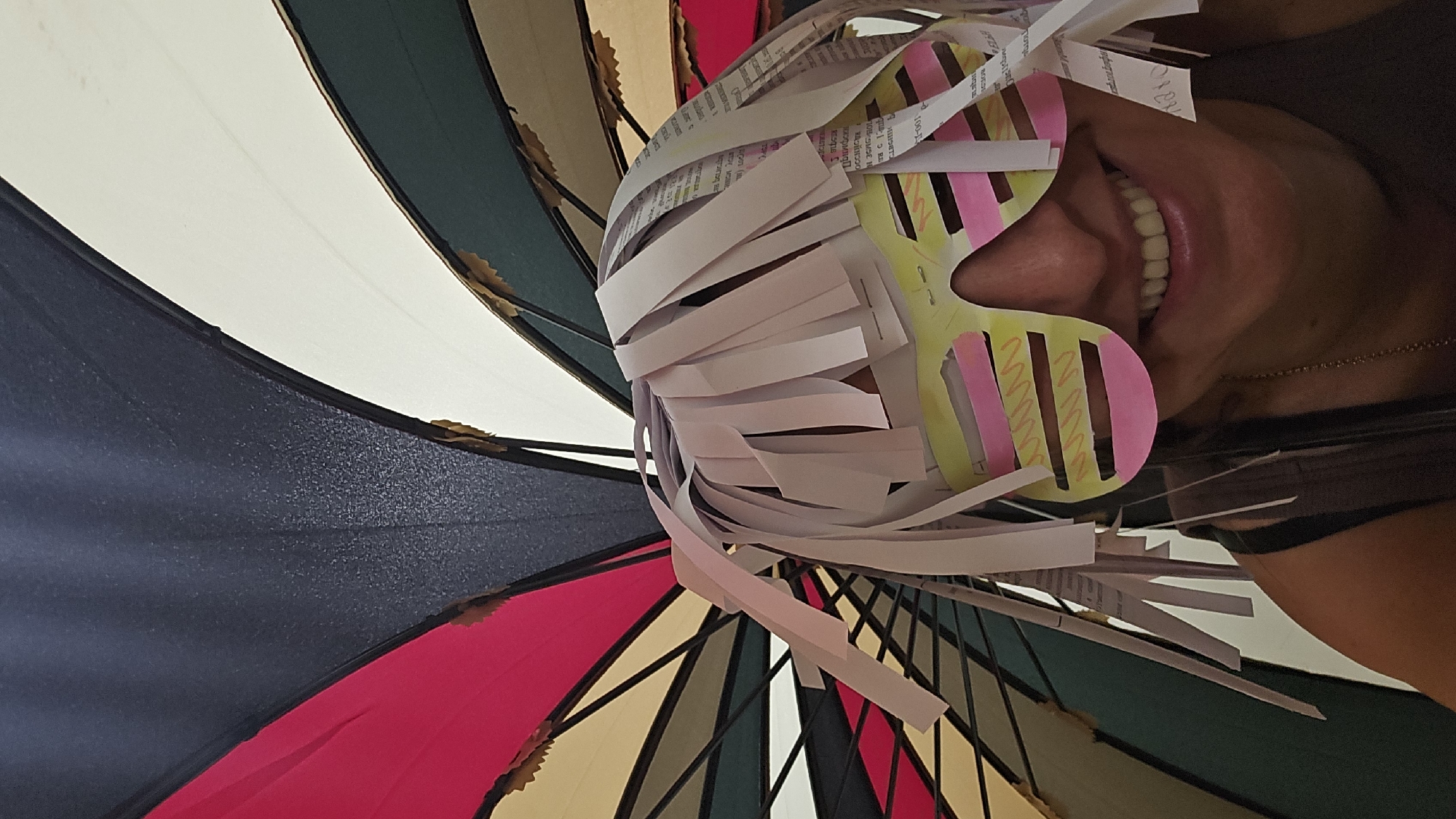 The photograph depicts a person's face wearing handmade paper sunglasses and a paper fringe attached to their head, viewed from a low angle looking upwards. The background consists of the interior of a multi-colored umbrella, featuring panels of black, red, beige, green, and white. The person is smiling, revealing their teeth. The location is unspecified.