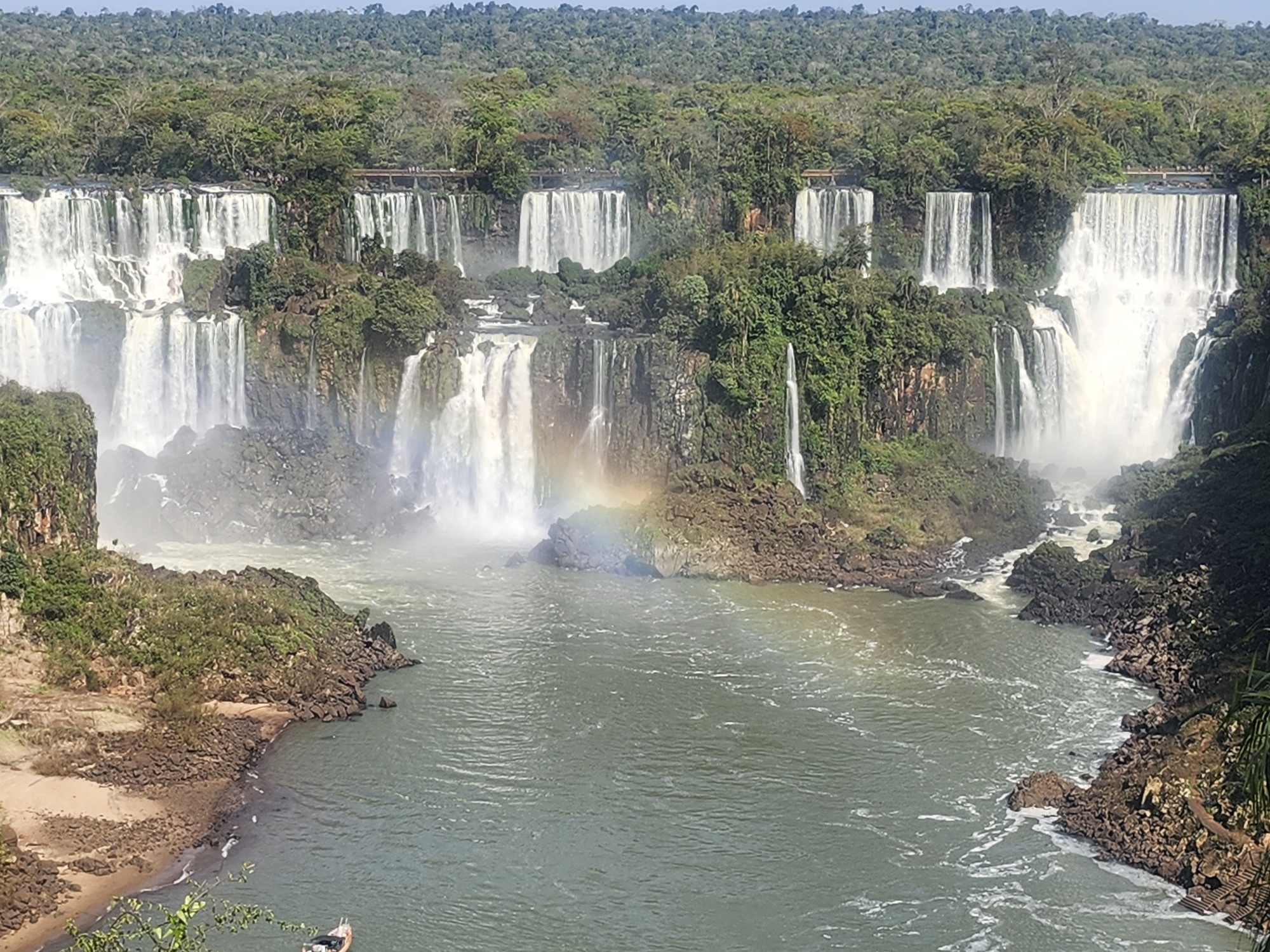 The image shows a panoramic view of Iguazu Falls, located on the border of Brazil and Argentina, near the city of Foz do Iguaçu, Brazil. The falls are a system of hundreds of individual waterfalls, stretching for about 2.7 kilometers. In this particular view, multiple cascades of white water plunge over rocky cliffs, creating a misty spray. Lush green vegetation covers the surrounding cliffs and the horizon is dominated by a dense forest. A river flows in the foreground, its surface rippled by the powerful currents. A faint rainbow is visible in the mist of one of the waterfalls, indicating sunlight interacting with the spray. Small wooden walkways and viewing platforms are visible along the top of some of the falls, with a few people standing on them, observing the scene. A small boat is anchored near a sandy shore on the left side of the river.