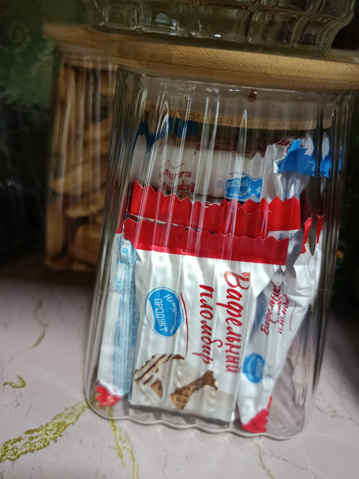 A clear glass jar with a wooden lid is filled with individually wrapped cookies. The cookies are silver and red with blue lettering that reads "ПРОДУКТ" which translates to "PRODUCT". Some of the cookies have images of wafers. The jar is placed on a light-colored, speckled surface, possibly a table, with a green, abstract pattern in the background. This scene appears to be an indoor setting.