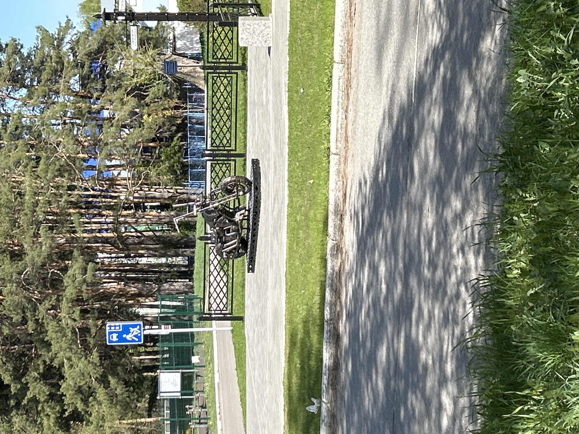 The image depicts an outdoor scene with a paved walkway and a road running parallel. Lush green grass borders both the walkway and the road. A metallic sculpture of a motorcycle is positioned on the walkway, enclosed within a black, ornate metal fence. Behind the fence and walkway, a dense forest of pine trees is visible. A blue road sign with white symbols is also visible near the trees. The shadows cast on the road suggest a sunny day. The location is Chaikovsky, Russia.