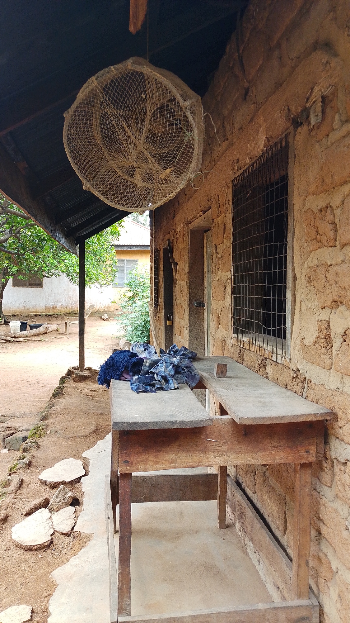 This image captures an outdoor scene in Donga, Nigeria, likely in a rural setting. The focus is on the exterior of a building constructed with rough, earth-toned materials, possibly mud or adobe bricks, which shows signs of age and weathering. A corrugated metal roof extends over a sheltered area in front of the building, supported by a wooden post.
Hanging from the roof is a large, circular object made of woven material, resembling a trap or a basket, possibly for fishing or storing something. In front of the building, on a simple, wooden table or bench, are piles of folded fabric, appearing to be clothing, one piece a dark blue, the other a checkered pattern of blues and grays. A small, light-colored wooden block sits on the table near the clothing.
The building features a window with a metal grate, suggesting a need for security. To the left of the building, a glimpse of another structure with a lighter colored wall and a metal roof is visible, along with lush green trees and vegetation. The ground is a mixture of dirt and concrete, with scattered stones and a small concrete path. The overall impression is one of a simple, functional living space, possibly in a village or a compound. There is no visible text in the image. The lighting suggests it is daytime, with no specific indicators of time of day or weather, other than the open-air setting.