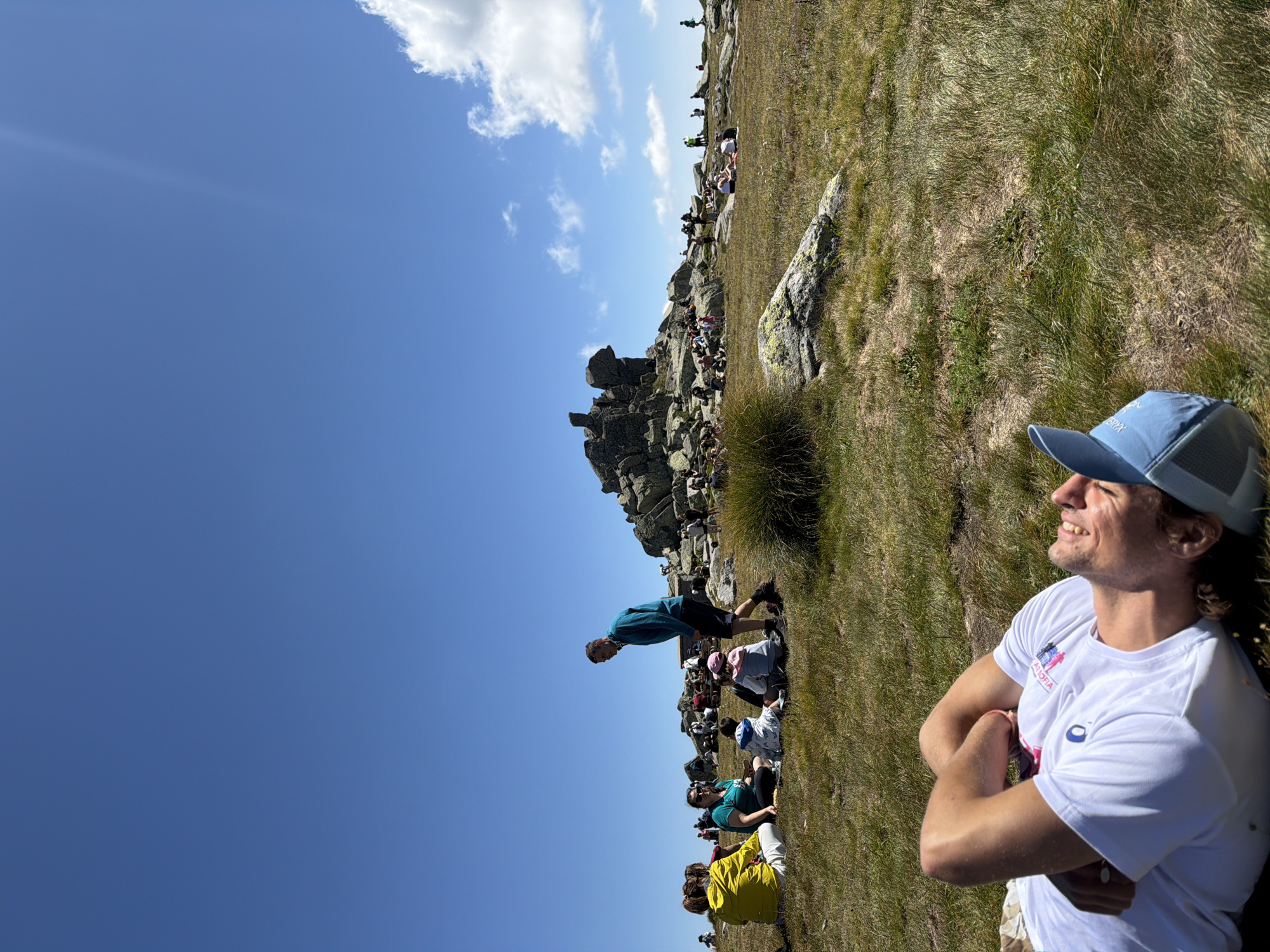 In a mountainous outdoor setting, under a clear blue sky with a few scattered clouds, a group of people are gathered on a grassy slope dotted with rocks. In the foreground, a young man is lying on his back in the grass, resting with his arms behind his head and smiling. He is wearing a white t-shirt and a blue baseball cap. Further back, several individuals are seated and standing on the grass and among the rocks. A man in a teal long-sleeved shirt and dark shorts is standing and looking towards the left. Other people are seen in smaller groups, some seated on blankets or directly on the ground, engaged in conversation or simply enjoying the scenery. Behind the people, a large rock formation dominates the midground, with a few small structures visible near its base. The terrain is covered in dry grass and scattered rocks, indicating a high-altitude, possibly arid environment. The scene suggests a recreational gathering, possibly a picnic or hike, taking place on a sunny day. The location is identified as the city of Bistritsa, Bulgaria, likely a mountainous area within or near the city.
