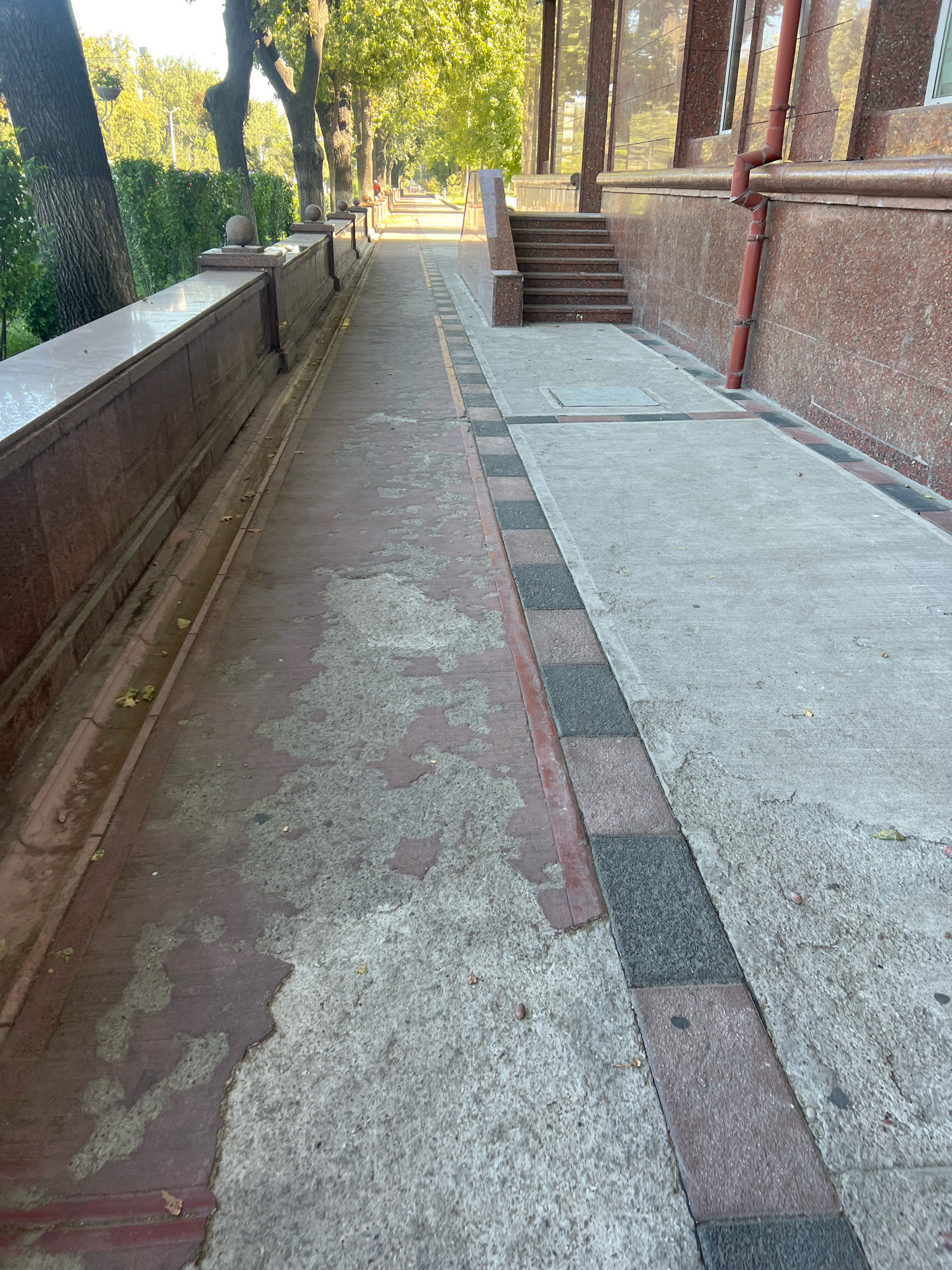 The image shows a perspective view of a pathway in an urban environment. The pathway is divided into two sections. The left section is made of reddish-brown paving stones with areas of discoloration and wear, suggesting it is older or has been exposed to water. It is bordered by a red granite wall with decorative finials and a row of trees with lush green foliage. A drainage channel runs along the base of the wall.
The right section of the pathway is made of grey concrete. It is bordered by a decorative strip of alternating red and dark grey paving stones. Further to the right, a building facade is visible, clad in red granite. A dark red downspout runs vertically down the facade. A set of concrete stairs leads up to an entrance on the right side of the image. The overall scene suggests a well-maintained public space, possibly a park or a walkway alongside a public building. The lighting indicates it is daytime, with sunlight casting shadows.
