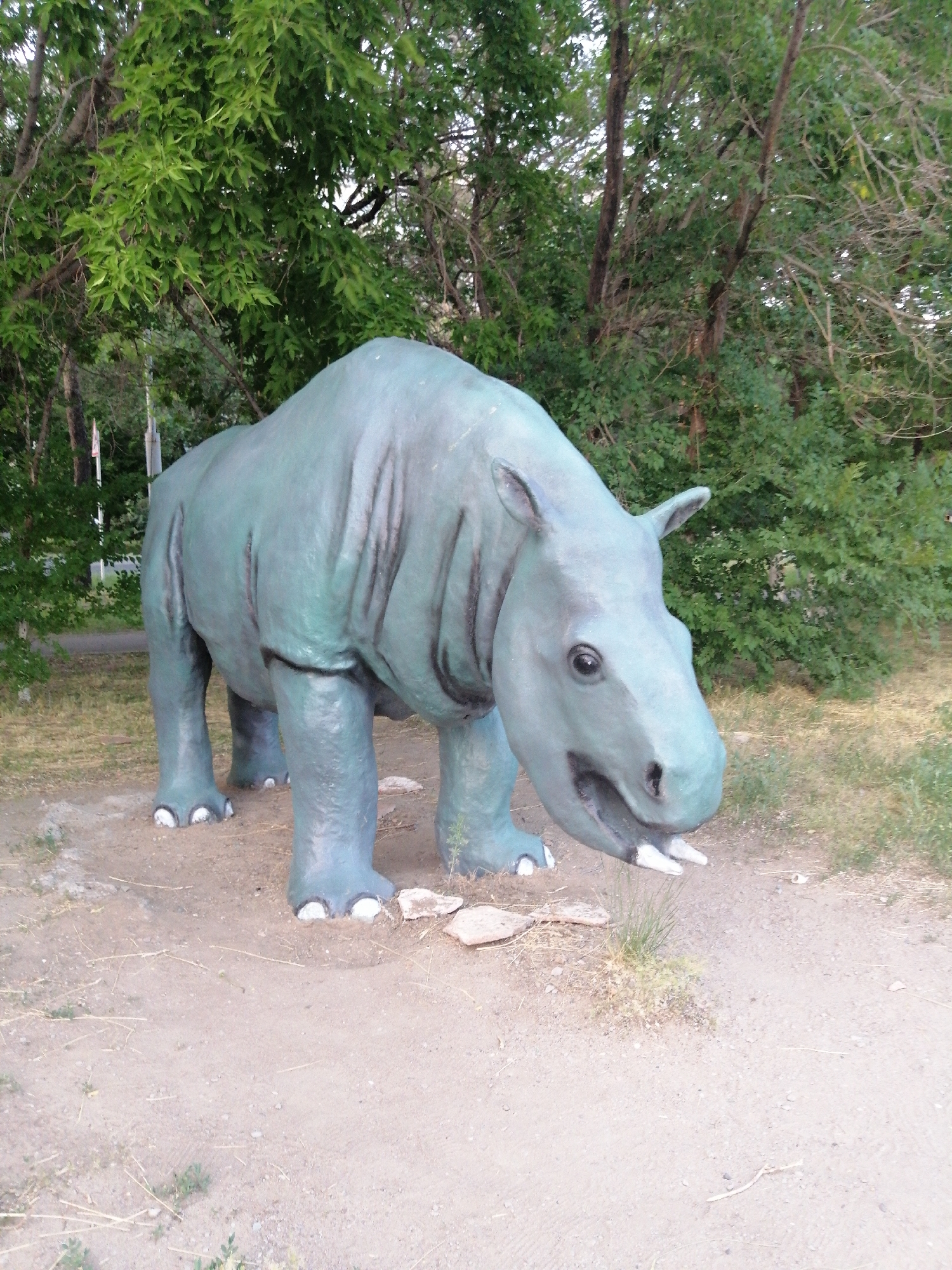 This media file features a large, teal-colored statue of a prehistoric mammal, likely an Indricotherium, positioned outdoors. The statue is life-sized and detailed, with wrinkled skin, four stout legs, and a prominent head with small ears and visible tusks. The creature's mouth is open, revealing more of its tusks and a dark interior. The statue stands on a dirt path, surrounded by dry grass and small stones.
In the background, a dense forest of green trees provides a natural backdrop, with their leaves creating a dappled effect of light and shadow. The foliage is lush, suggesting a temperate climate and possibly a park or natural reserve setting. The lighting indicates it is daytime, with no harsh shadows suggesting it is not midday. The overall atmosphere is serene and educational, likely part of an outdoor museum or paleontological exhibit. No people are visible in the frame, and there is no discernible text. The setting appears to be a tranquil, natural environment, likely in or near Pavlodar, Kazakhstan, given the provided location context.