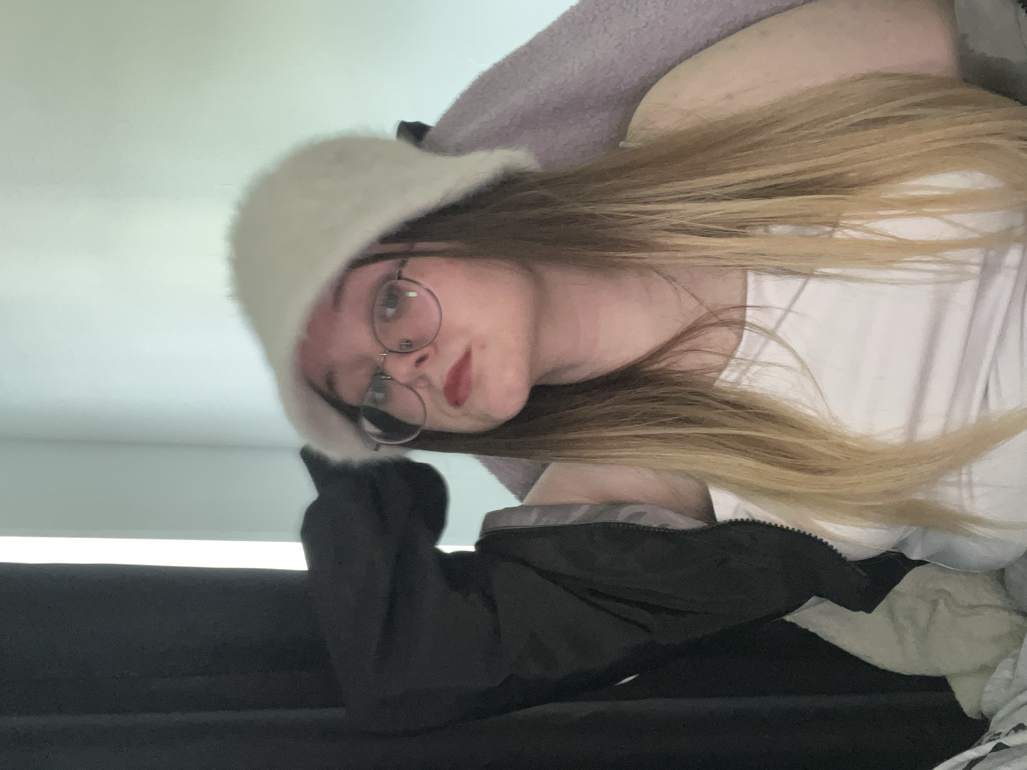 A young person with long, light brown hair is wearing a white, furry hat and round-framed glasses. They have fair skin and red lipstick. They are wearing a white shirt under a black jacket. The person is sitting on a black surface, possibly a couch. A grey or lavender blanket is partially visible in the background. The location is not identifiable as Pushchino, Russia. The image is oriented sideways.