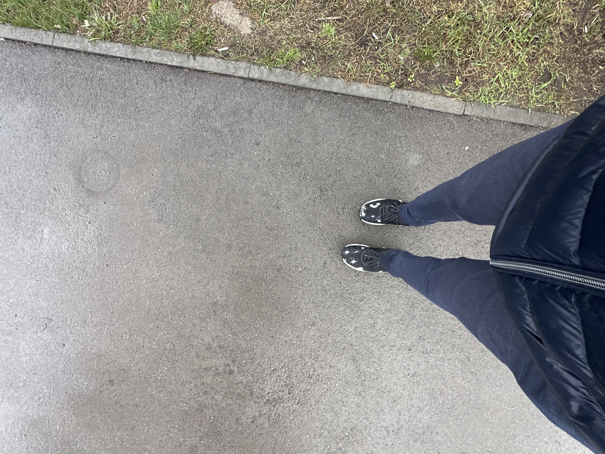 The image shows a person standing on a grey concrete surface, likely a sidewalk or pavement. The person is wearing dark denim pants and black athletic shoes with white soles and laces. The right side of the frame captures a portion of a dark blue, puffy jacket. Adjacent to the pavement is a strip of land with short green grass and some bare patches of dirt. A thin concrete curb separates the pavement from the grassy area. A circular discoloration is visible on the left side of the pavement.