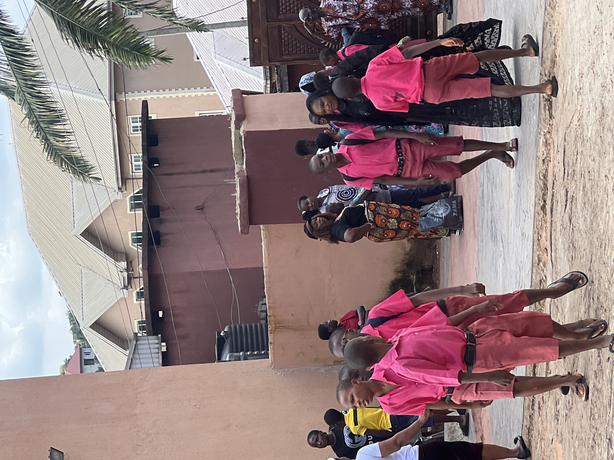 The image depicts an outdoor scene in the city of Nnewi, Nigeria, likely on a street or within a school compound. In the foreground, several young boys are walking, all wearing matching pink short-sleeved shirts and coral-colored shorts. Most of the boys are wearing black belts. Some have backpacks. To the left, an adult woman is partially visible, wearing a white t-shirt and dark shorts. Behind the boys, a group of adults and children are gathered. The architecture in the background suggests a residential or institutional setting, with buildings featuring corrugated metal roofs and decorative facades. There are palm trees and wires visible, indicating a tropical or sub-tropical environment. The ground appears to be paved with concrete and dirt. The overall atmosphere seems to be one of daily activity, possibly during a school day or an event.