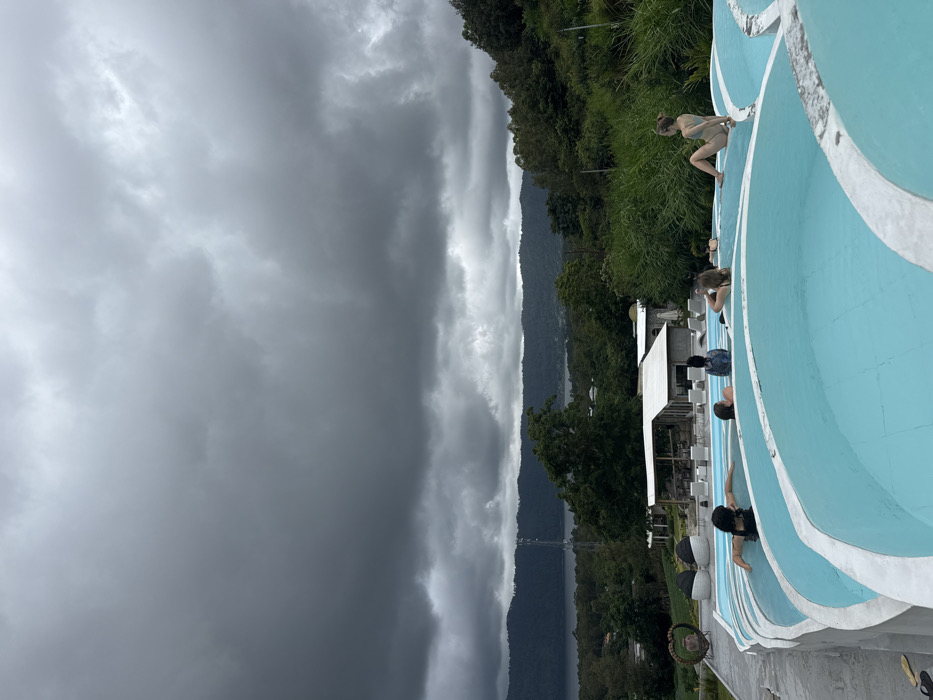 The image displays an outdoor swimming pool complex in Toyabungkah, Indonesia, situated on a slope overlooking Lake Batur and surrounding forested mountains under a heavily overcast, dark gray sky. In the foreground, a multi-tiered series of light blue pools with white, undulating edges extends across the bottom of the frame. Approximately seven individuals are visible. One female, wearing a light-colored swimsuit, sits on the edge of the uppermost pool tier on the right, facing left with her legs drawn up. Another female, with dark hair, lies prone in a lower pool section on the left. Five other individuals are partially submerged within the various pool tiers, appearing to be in relaxed positions. In the midground, several white-roofed buildings, likely part of a resort, are visible among dense green foliage. A tall, slender cellular tower stands amidst the trees to the left of the central view. The background features the expansive, dark surface of Lake Batur, bordered by uniformly dark green, tree-covered mountains. Two flip-flops are visible on the ground to the left of the leftmost pool.