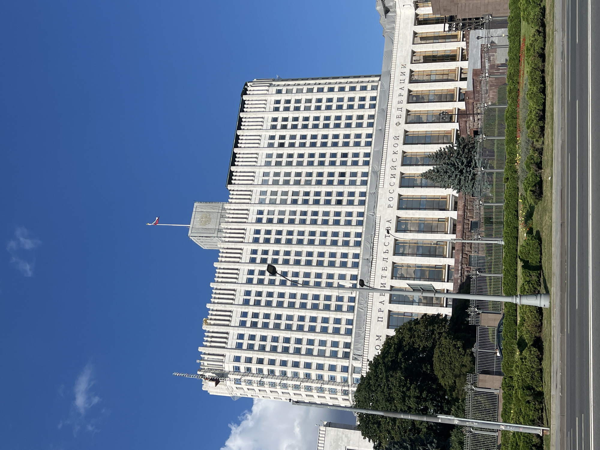 The image shows a large, white, neoclassical building with many windows, set against a bright blue sky with a few white clouds. The building has a prominent inscription in Russian that reads "ДОМ ПРАВИТЕЛЬСТВА РОССИЙСКОЙ ФЕДЕРАЦИИ," translating to "HOUSE OF THE GOVERNMENT OF THE RUSSIAN FEDERATION." A Russian flag flies from a flagpole on the roof. In front of the building is a metal fence, a manicured garden with bushes and some flowering plants, and a street with road markings. Two lampposts are visible in the foreground. Trees, including a large dark green fir tree, are present in the garden area. A dark-colored car is visible through the fence. The building appears to be under some renovation or construction, as a temporary structure with a lift mechanism is attached to the side of the building. This building is the White House of Russia, located on Krasnopresnenskaya Embankment in Moscow.