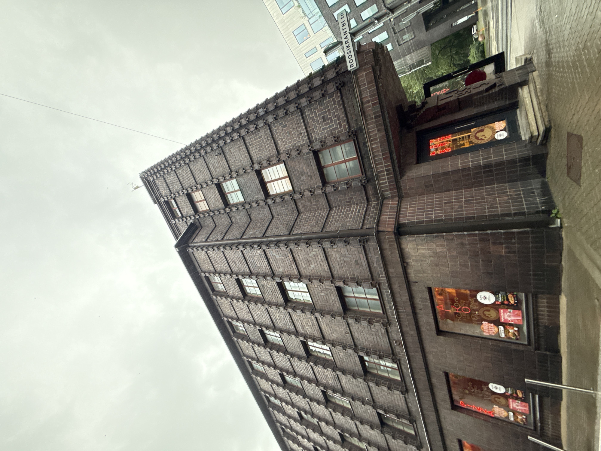 The image shows a wide-angle view of a dark brick building with a unique, stepped facade and numerous windows. The building has a sharp corner that angles upwards. The windows on the lower level of the building display advertisements for "Pizza" and "Kohvik" (cafe in Estonian), with neon signage visible on the windows and doors. A street sign in the upper right corner reads "ROOSIKRANTSI tn". Adjacent to the brick building, a modern, lighter-colored building is visible. The sky is overcast, and the ground appears wet, suggesting it may have recently rained or is currently raining. The image appears to have been taken from street level, looking up at the buildings.
