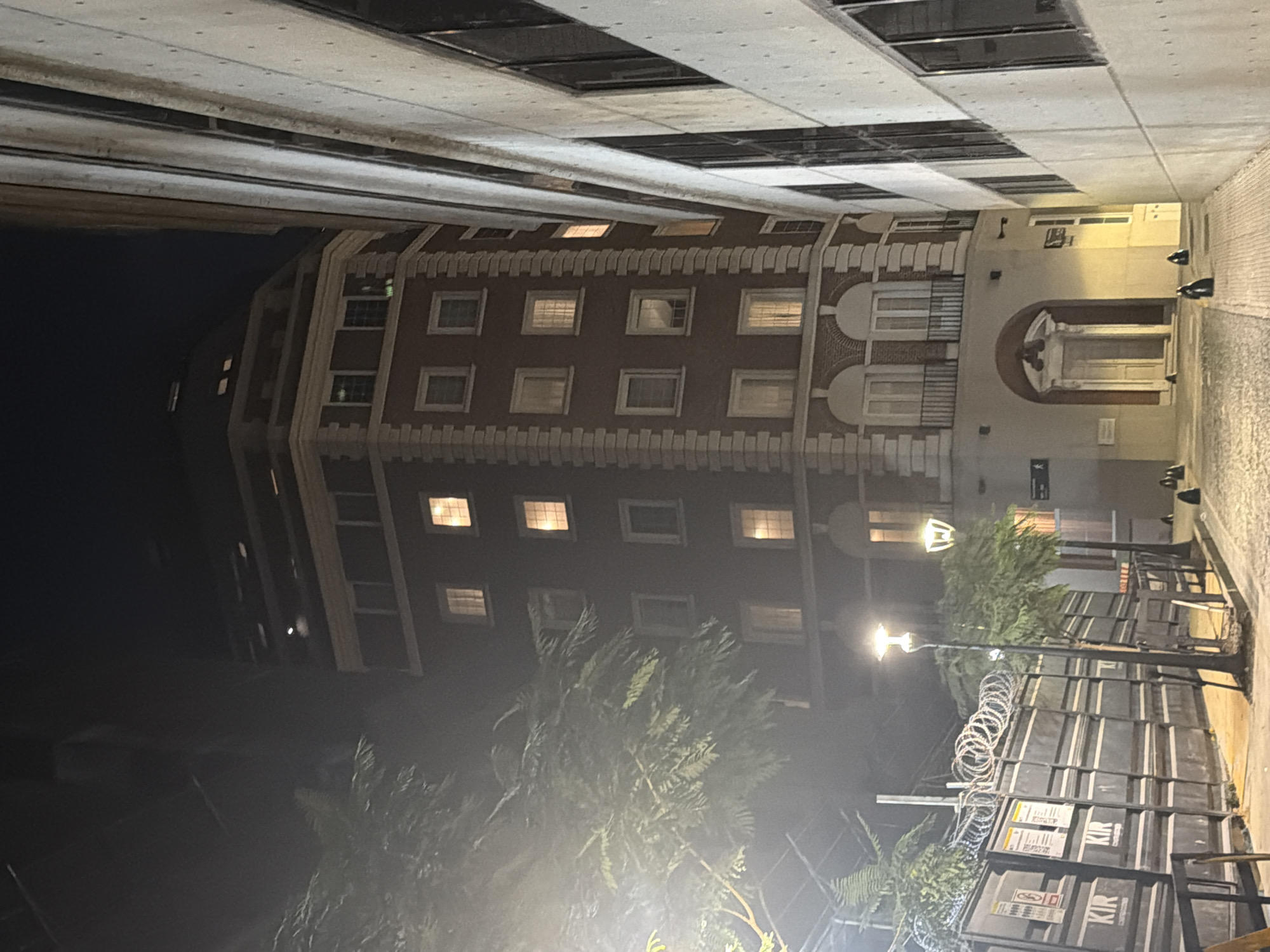 The image shows a building in Buenos Aires, Argentina at night. The building has a brick facade and numerous windows, some of which are illuminated. A decorative archway is visible on the right side of the building. In the foreground, there are trees, streetlights, and a barricade with razor wire. The street is paved and wet, suggesting recent rain. Additional building overhangs the sidewalk.