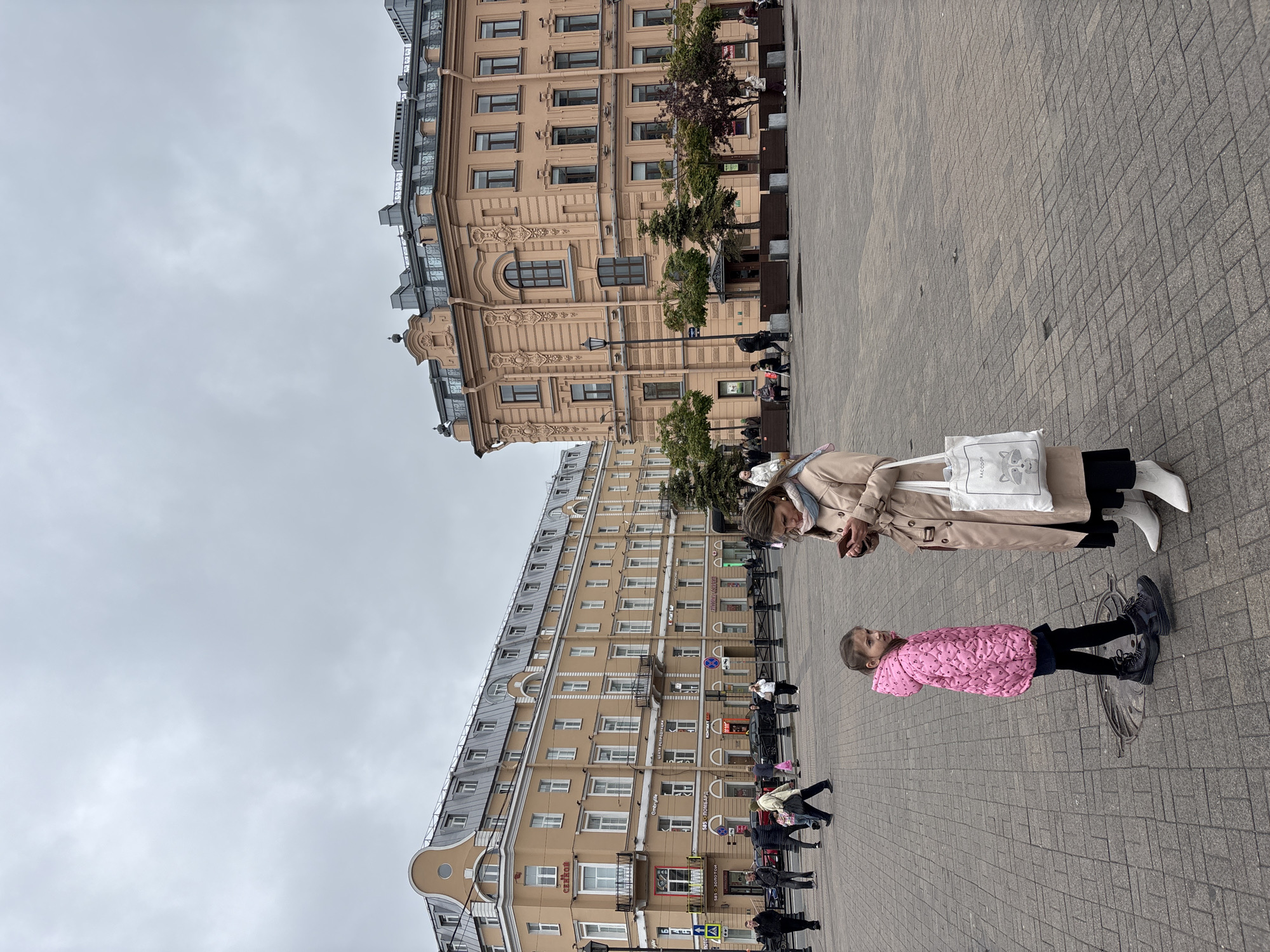 The image shows a scene in Saint Petersburg, Russia. The background features two large buildings with a light tan color and multiple windows, characteristic of the city's architecture. The sky is overcast. The foreground is a pedestrian square paved with gray bricks. On the square, there is an adult woman and a child. The woman is wearing a beige trench coat, carrying a white bag, and using a smartphone. The child is wearing a pink quilted jacket, black leggings, and boots. They appear to be interacting, possibly talking or looking at something on the woman's phone. Several other people are walking in the background near the buildings.