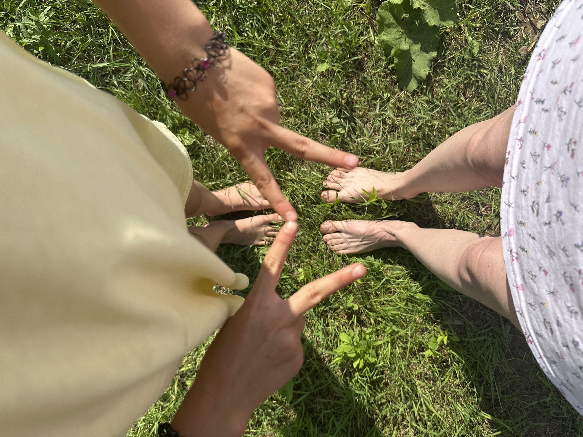 A first-person perspective shot shows bare feet standing on green grass. Two individuals' hands form a heart shape above the feet. One person wears a light yellow garment, and another wears a white garment with a floral pattern. A dark bracelet with pink accents adorns one wrist. The scene is outdoors, with sunlight casting shadows. No specific city landmarks or identifiable structures are visible in Mókino, Russia.