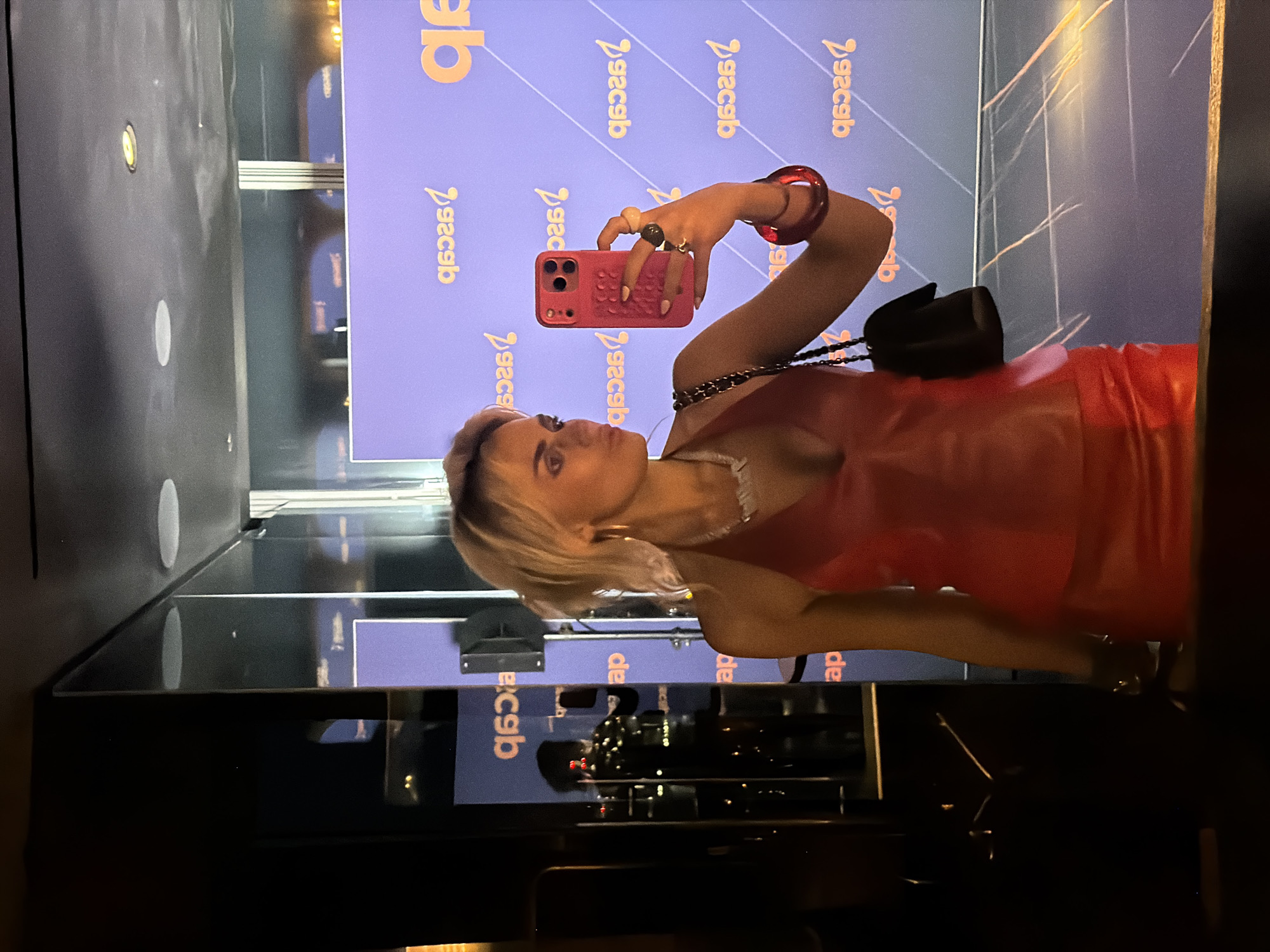 A woman with blonde hair, wearing a red halter top and skirt, takes a selfie in a mirror. She holds a pink smartphone in her right hand, and her left hand rests on her hip. She is adorned with a necklace and several rings. Behind her, a purple backdrop displays the reversed text "ASCAP". The scene appears to be in a dimly lit interior, possibly a venue in London, United Kingdom.