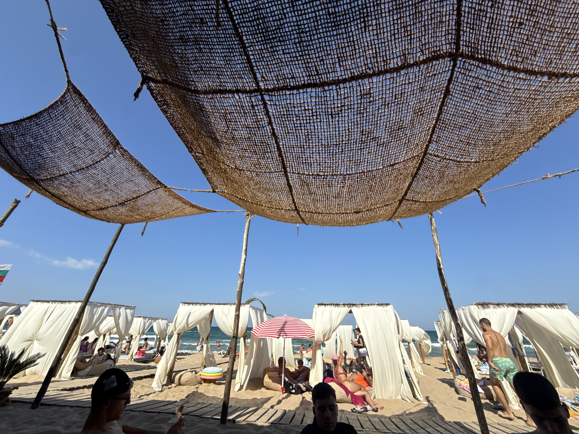 The image depicts a sunny day at a beach in Primorsko, Bulgaria. In the foreground, a woven canopy made of natural fibers is supported by wooden poles, casting shadows on the sand. Beneath the canopy, a row of white cabanas with draped fabric creates shaded seating areas for beachgoers. People are seen relaxing in the cabanas, some lying on beanbag chairs, while others are engaged in conversation or resting. Further back, the turquoise waters of the Black Sea are visible, with small waves and a few distant figures enjoying the ocean. A red and white striped umbrella stands out among the cabanas, adding a splash of color to the scene. The beach is sandy, with wooden walkways leading between the cabanas. The sky is clear blue, indicative of fair weather. The overall atmosphere is one of leisure and relaxation on a popular beach destination.