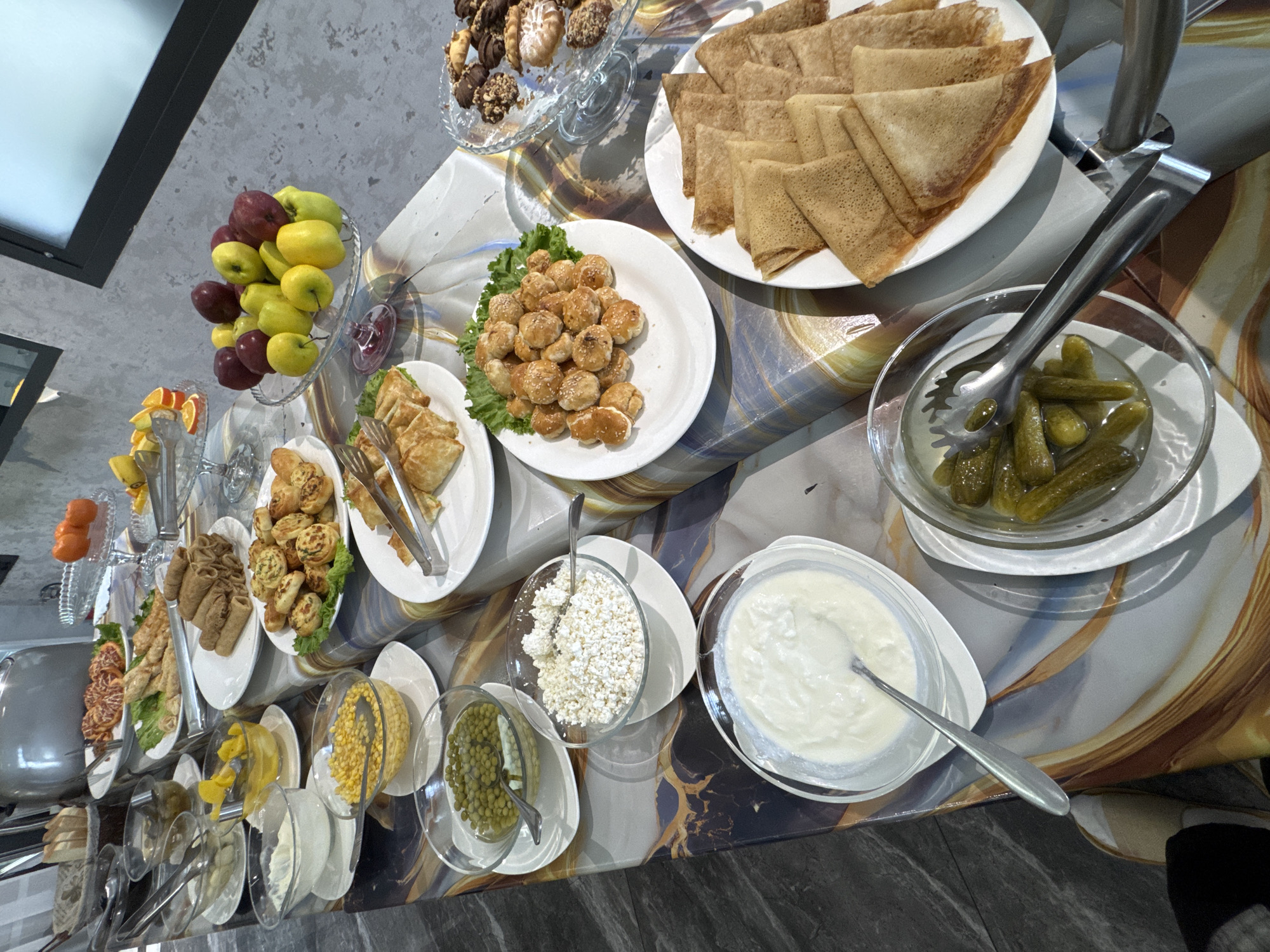 The image depicts a buffet-style spread of various foods, likely in a hotel or restaurant in Tashkent, Uzbekistan.
**Subjects & Objects:**
* **Food Items:** Plates and bowls contain a variety of foods including:
* Folded pancakes or crepes
* Small fried or baked dough balls, possibly "baursaki" or similar
* Pickled cucumbers
* Yogurt
* Cottage cheese
* Canned corn
* Canned green peas
* Assorted pastries
* Rolled savory pancakes
* Sliced fruits (apples, oranges, etc.)
* Cookies and/or small cakes
* **Tableware:** Plates, bowls (some on stands), serving spoons, and tongs are present.
* **Table:** The food is arranged on a long table covered with a patterned tablecloth.
**Environment:**
* The setting is likely indoors, possibly a hotel dining room or a restaurant.
**Actions & Interactions:**
* The scene is static, showing food prepared for a meal or event.
**People:**
* While there are no people visible in the immediate frame, the arrangement of food indicates preparation for a meal that would be consumed by people.