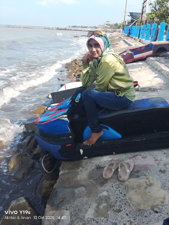 An Indonesian woman is seated barefoot on a modified personal watercraft, likely a repurposed jet ski, at a coastal location in Limbangan, Indonesia. She wears a light green long-sleeved shirt, dark blue jeans, and a multi-patterned hijab, with dark sunglasses perched on her head. Her left hand is positioned near her chin, and she displays a subtle smile towards the camera. The watercraft, predominantly blue and black with red and pink accents, rests on a concrete platform at the edge of a rocky shoreline. Murky water with moderate waves crashes against the rocks. A pair of dirty, light-colored slip-on sandals are on the concrete platform to the viewer's left. In the background, a blue-railed barrier and a partially overturned wooden boat are visible on the concrete. A rocky breakwater extends into the sea under a clear sky with scattered clouds. The scene is illuminated by bright daylight. An on-screen watermark indicates the image was captured on October 12, 2025, at 14:47.