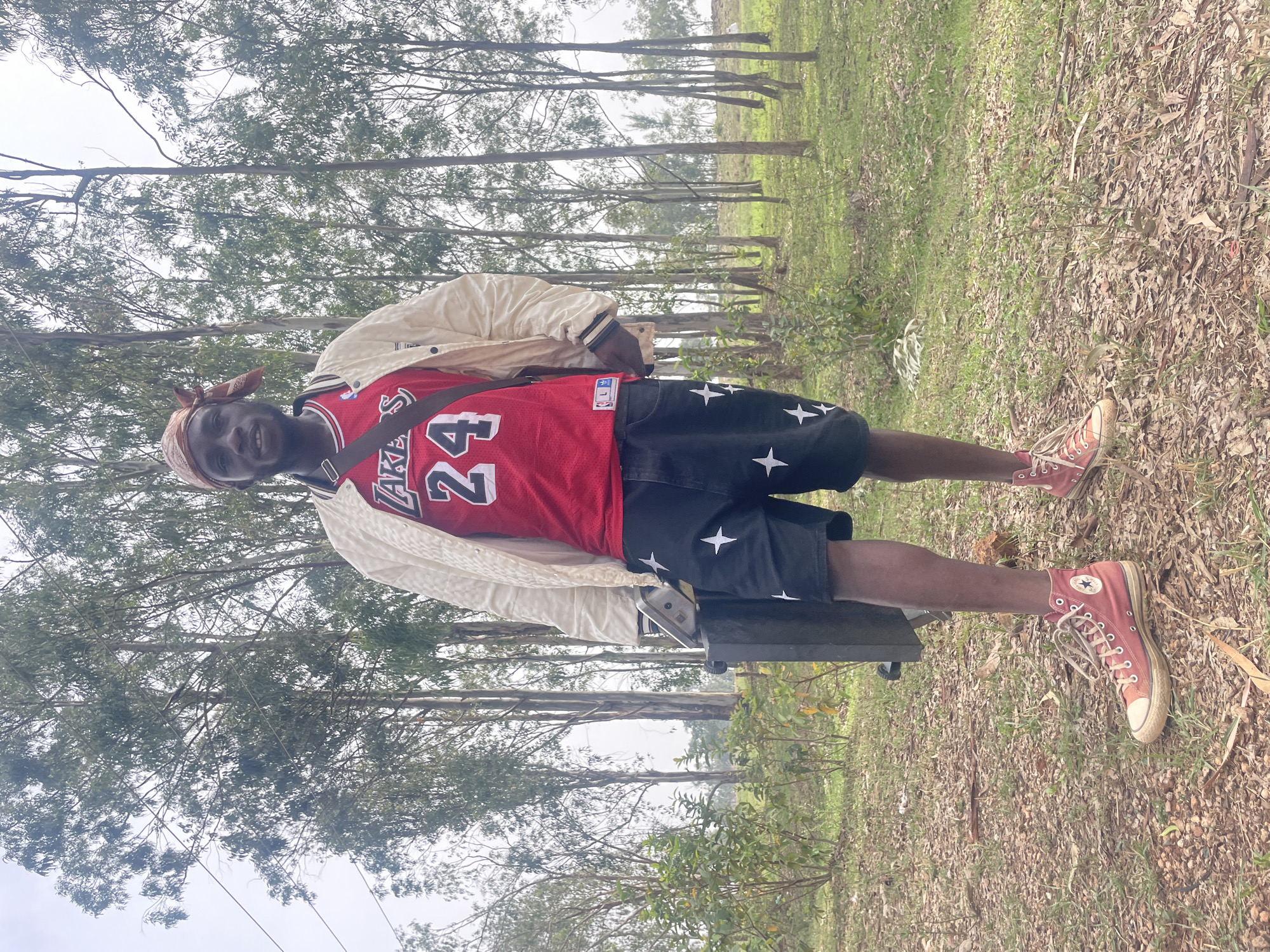 A young man stands outdoors. He wears a red Lakers jersey with the number 24, black shorts with white star patterns, and red high-top sneakers. A beige jacket is draped over his shoulders. He has a brown bandana on his head and is making a hand gesture. He carries a dark-colored bag across his body. The background shows trees and foliage, suggesting a park or wooded area. The ground is covered in leaves and grass. The picture is in a vertical format. The location is Durumi, Nigeria.