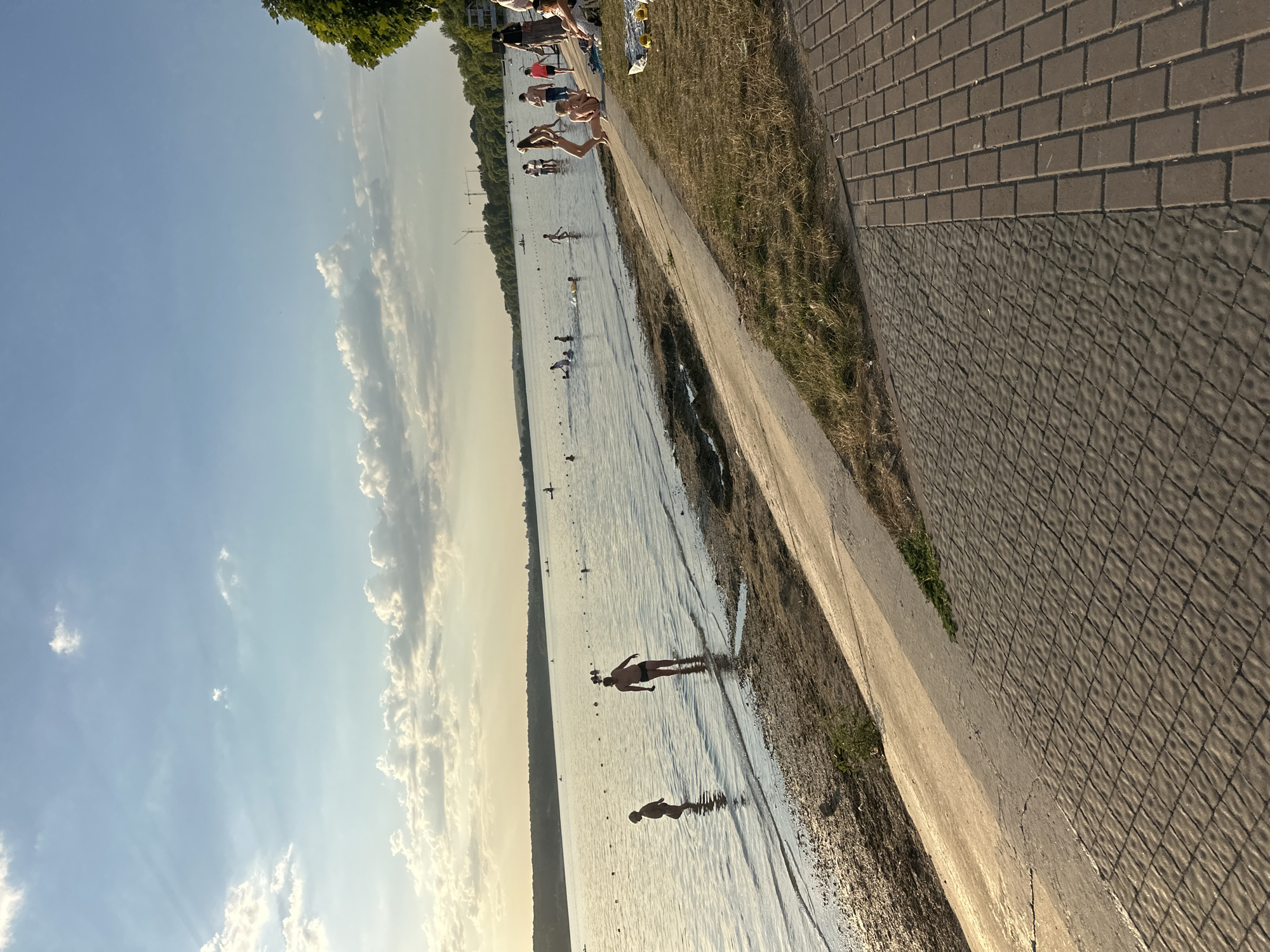 This is a wide shot of a lake or river during what appears to be late afternoon or early evening, possibly in Ternopil, Ukraine, given the context. The sky is a clear blue with scattered white clouds, some of which are illuminated by the setting sun. A treeline is visible on the far bank of the water, with some distant buildings and construction cranes suggesting an urban or semi-urban setting.
In the foreground, a paved walkway made of interlocking bricks runs diagonally across the image. Adjacent to the walkway is a sloped, grassy embankment that transitions to a sandy shore at the water's edge.
The water itself is populated with various individuals, many of whom are children and young adults swimming or wading. There are buoys marking an area in the water. On the sandy shore and grassy embankment, people are also seen sitting, relaxing, and observing. The overall atmosphere suggests a recreational scene, possibly a beach or park area along the waterfront. The lighting indicates a warm, pleasant day.