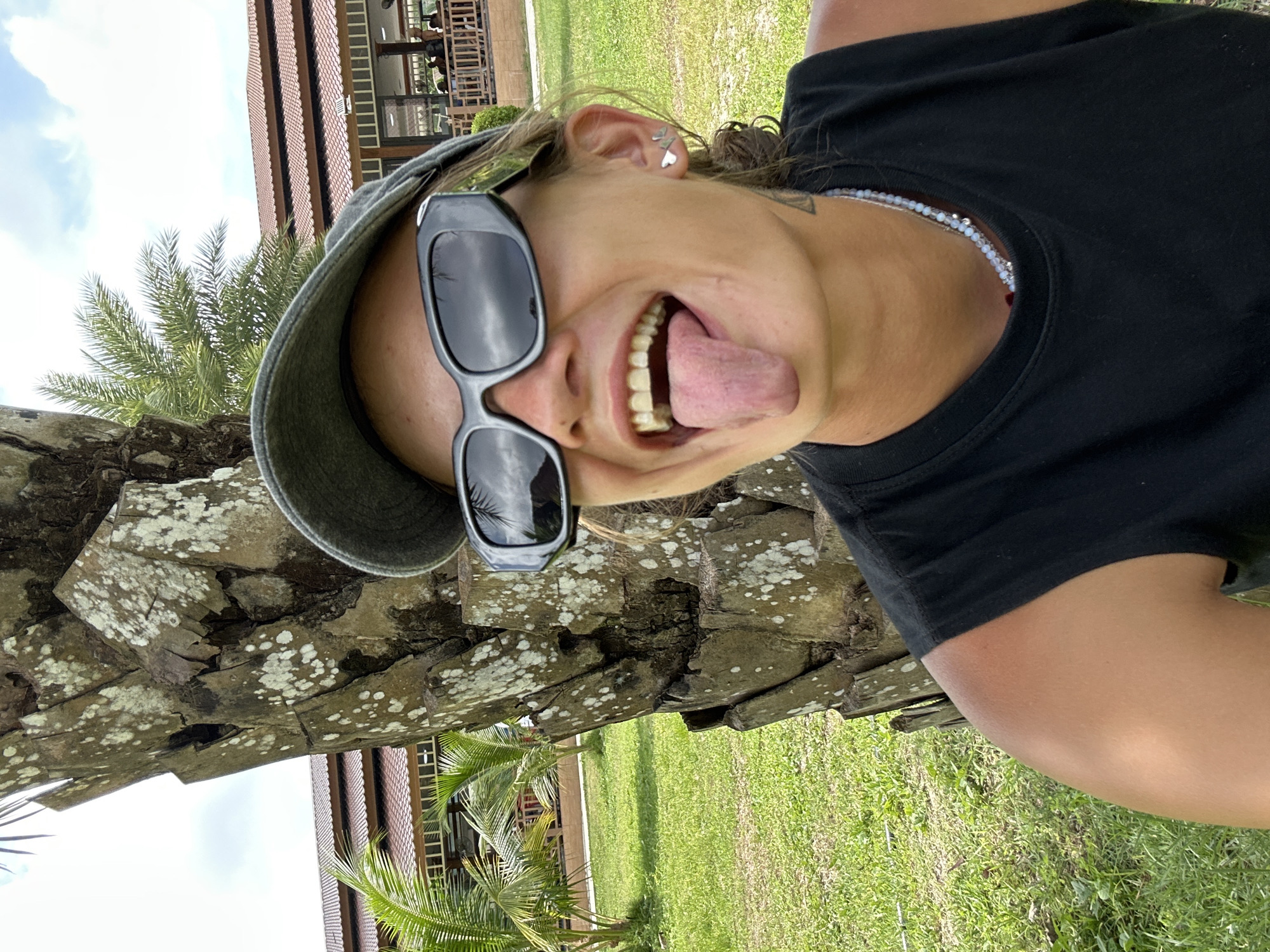 A person wearing sunglasses and a cap sticks their tongue out while leaning against a palm tree in Ao Nang, Thailand. The person is wearing a black tank top and a beaded necklace. In the background, there is a building with a red roof and palm trees, indicating a tropical environment. The sky is partly cloudy.