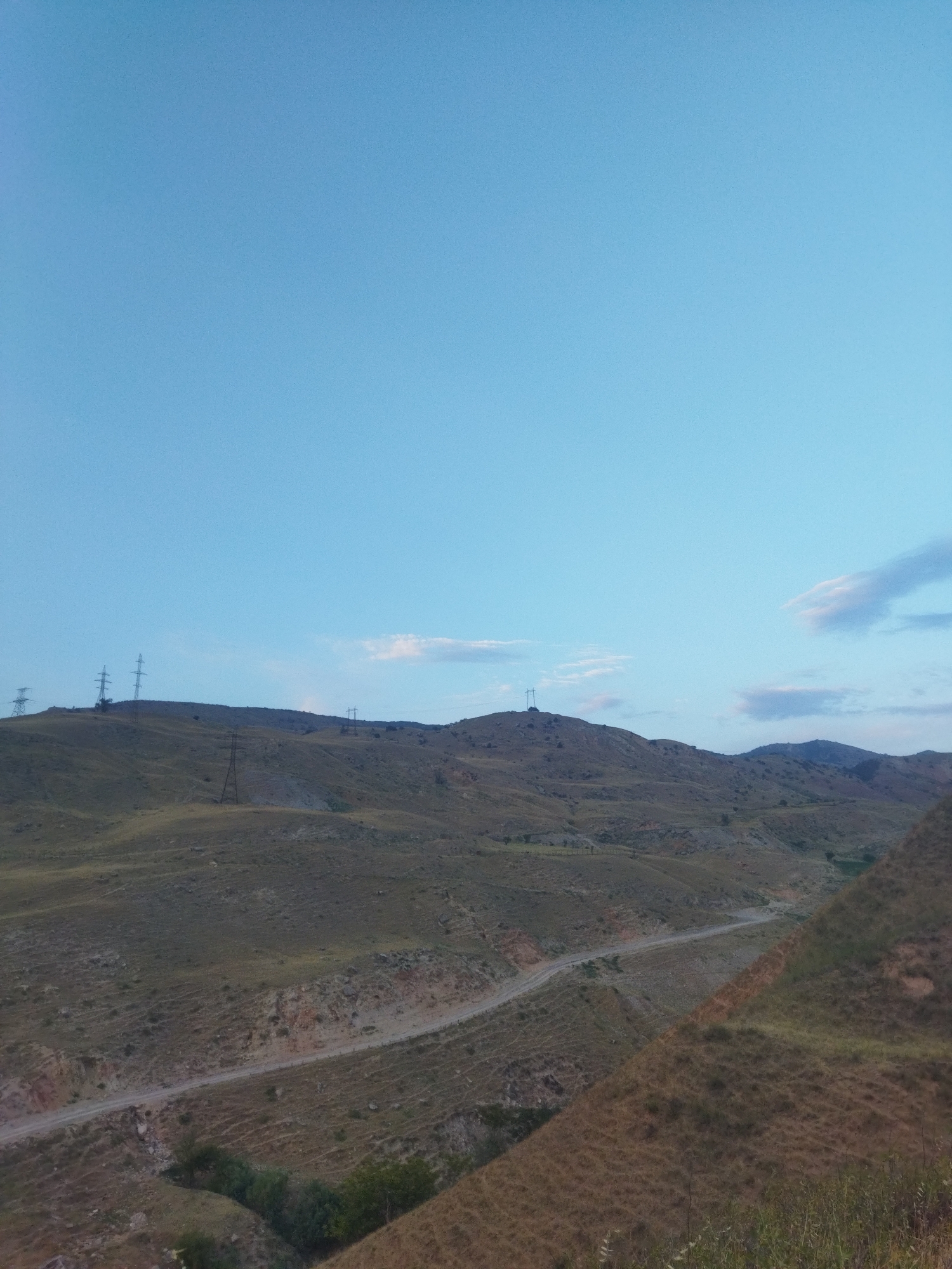 The image depicts a natural landscape with rolling hills and a clear blue sky. Power lines and transmission towers are visible on the horizon, suggesting the presence of infrastructure in the area. A dirt road winds through the hilly terrain, hinting at potential access routes or pathways within the landscape. The overall impression is one of a rural or semi-rural setting, likely in a region with a dry climate, given the appearance of the vegetation. The scene is devoid of any people or specific structures that would indicate a precise urban location within Mailuu-Suu City, Kyrgyzstan, but it shows a common type of landscape found in such regions.