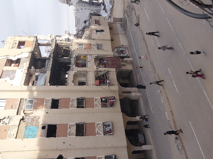 The image depicts a street scene in Khan Yunis, Palestinian Territory, dominated by a heavily damaged multi-story residential building on the left. The building's light beige facade shows numerous impact marks, bullet holes, and structural failures, with large sections of upper floors completely missing, exposing internal structures and rebar. Burnt areas are visible on the middle floors, and windows are either shattered or absent, some openings covered by tarps. Clothes are hanging on lines from several balconies, and a red patterned rug is draped over a balcony railing.
Below, a paved street with white lane markings spans the foreground. On the street and surrounding sidewalks, approximately 15-20 individuals are visible. Several children are walking and running, some appearing to play in groups. Adults are also present, walking on the street and sidewalks. One figure in a black abaya stands on the sidewalk near the damaged building, gesturing towards the right. Under an archway on the ground floor of the damaged building, a white car is parked, and two individuals are seated on the sidewalk. To the far right, beyond the street, further damaged structures and debris are visible, including the partially destroyed dome of what appears to be a mosque. Debris is scattered on the sidewalks and unpaved areas adjacent to the road.