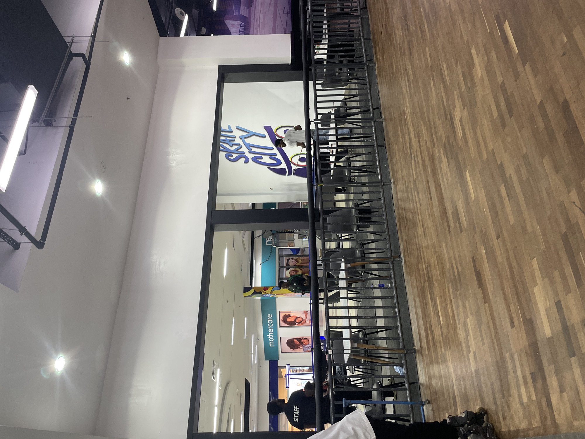 The image captures an indoor scene, likely a commercial establishment, with a prominent "SKATE CITY" mural on a white wall. The floor is made of polished wood planks. A dark metal railing separates the foreground from the area behind it, where tables and chairs are arranged. In the background, a "Mothercare" store is visible, suggesting a shopping mall environment.
Several individuals are present. To the left, a person wearing a black shirt with "STAFF" printed on the back stands facing away from the camera. To their right, another person in a green shirt and hat appears to be speaking with someone else. In front of the "SKATE CITY" mural, a person in a white t-shirt and jeans is interacting with the wall. The overall impression is that of a public space, possibly a recreational or retail area, within the city of Moba, Nigeria, though this specific location is not explicitly confirmed by any visual cues. The lighting appears to be artificial, with several light fixtures visible on the ceiling.