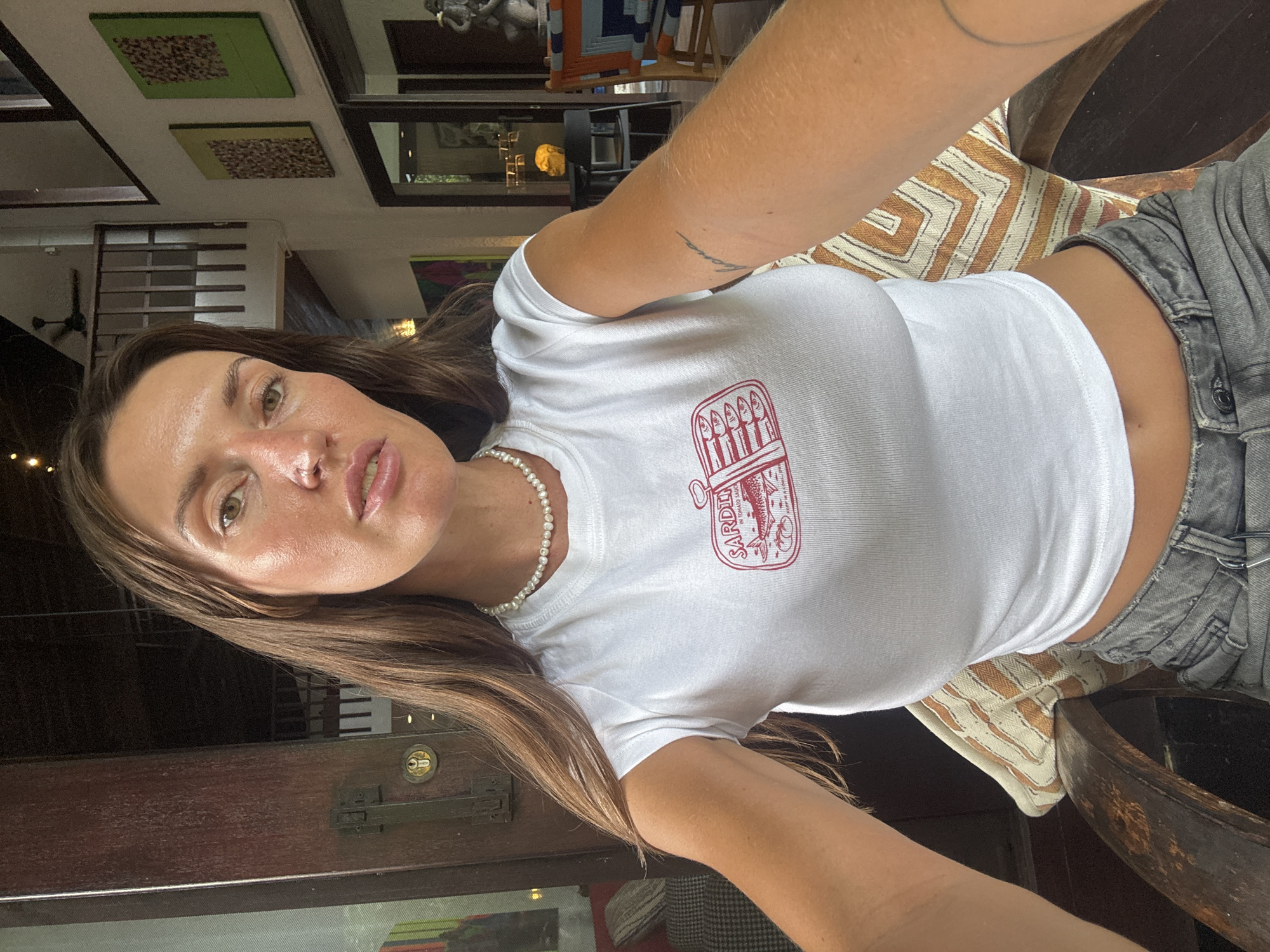 A woman with long brown hair sits in a wooden chair in a dimly lit room, possibly a restaurant or cafe, in Mengwi, Indonesia. She is wearing a white crop top with a red sardine can graphic and grey jeans. The room has high ceilings, exposed wooden beams, and is decorated with artwork on the walls. In the background, there is a staircase, tables, chairs, and glimpses of other patrons. The woman is looking directly at the camera, with a neutral expression.
